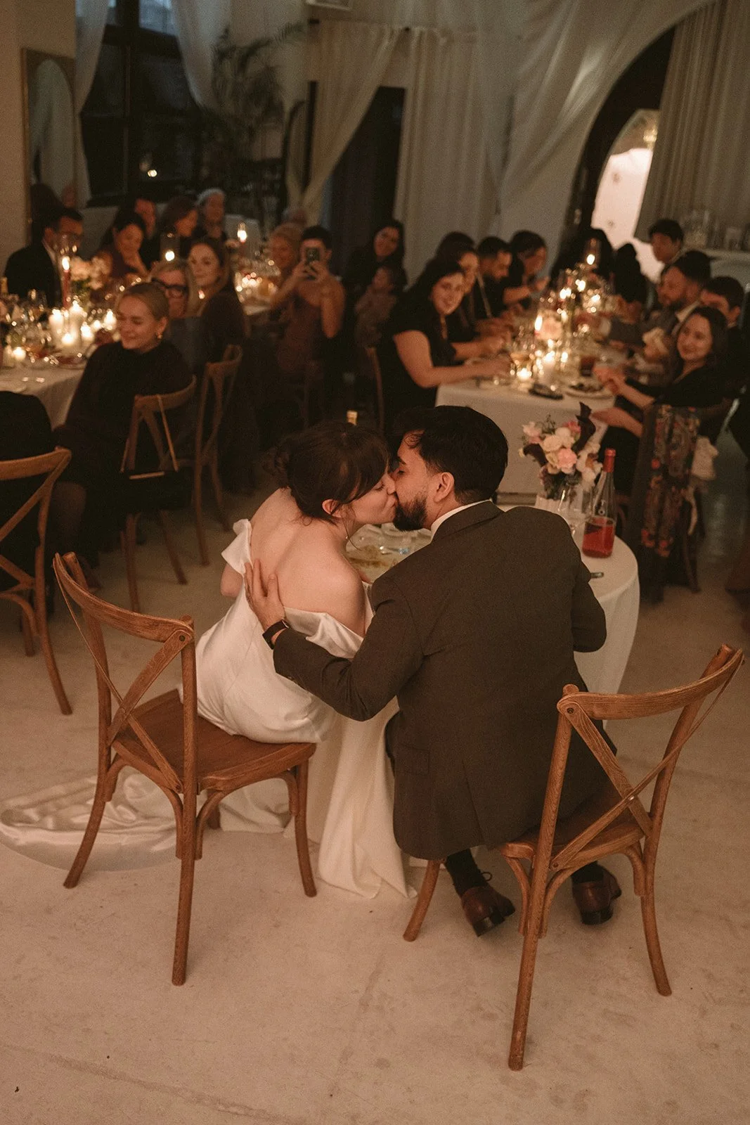 Couple kissing at wedding reception, seated on two wooden chairs, surrounded by guests at decorated dinner tables with candles and floral centerpieces.
