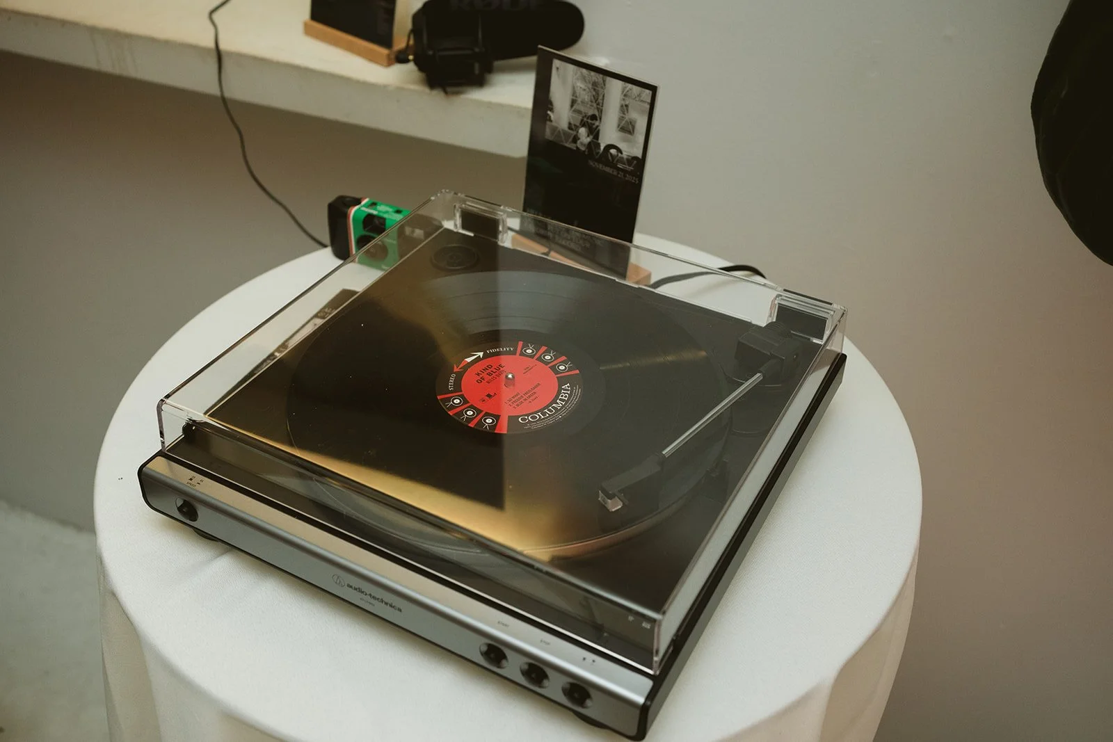 A turntable with a clear plastic cover on a white round table, with a red and black vinyl record intimate wedding. In the background, there's a camera, a small black box, and a photo display on the wall. marigolds florals nyc florist wedding florals 