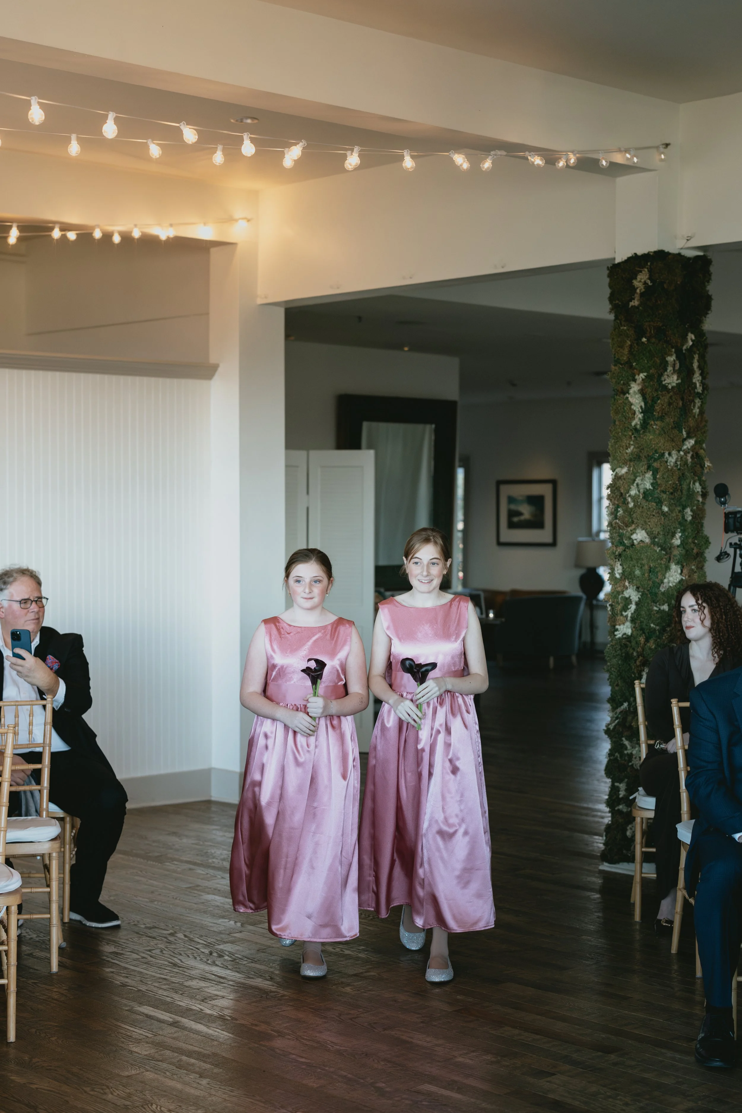 Two young girls in pink satin dresses walking down an aisle at a wedding, holding black calla lilies. The girls are smiling and wearing matching silver shoes. Guests are seated on either side, with some taking photos. The reception area has hanging s