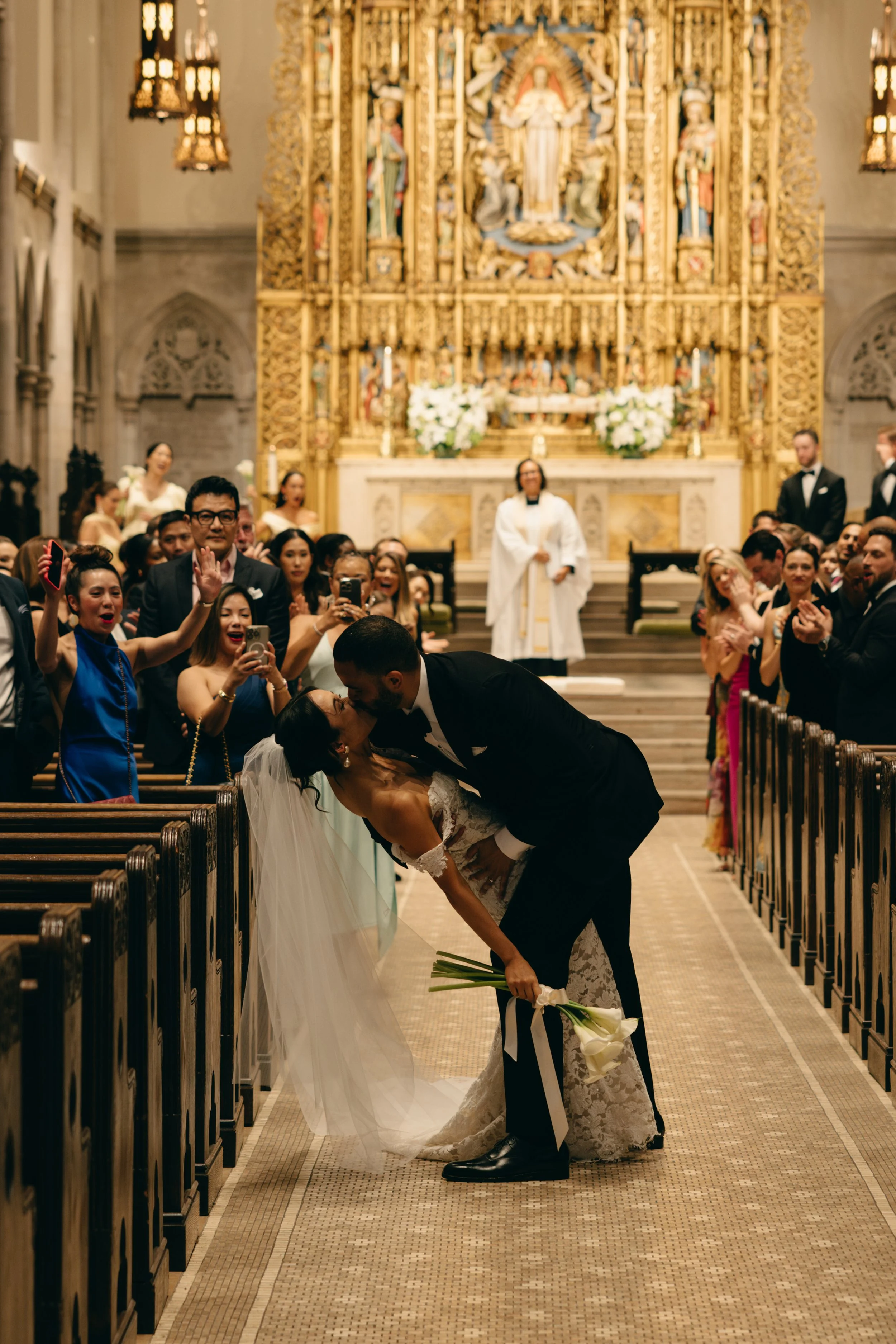 A newlywed couple kissing inside a church with guests watching and taking photos, during their wedding ceremony. marigolds florals nyc florist wedding florals central park boathouse wedding