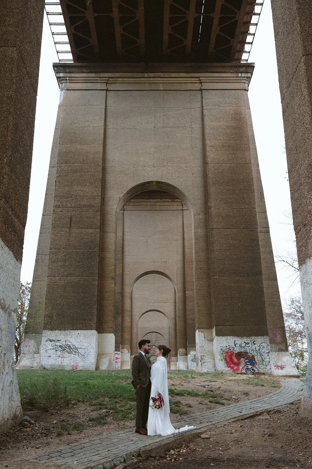 A bride and groom standing under a bridge, facing each other, with the bride holding a bouquet of flowers in her left hand. marigolds florals nyc florist wedding florals atmosfera wedding