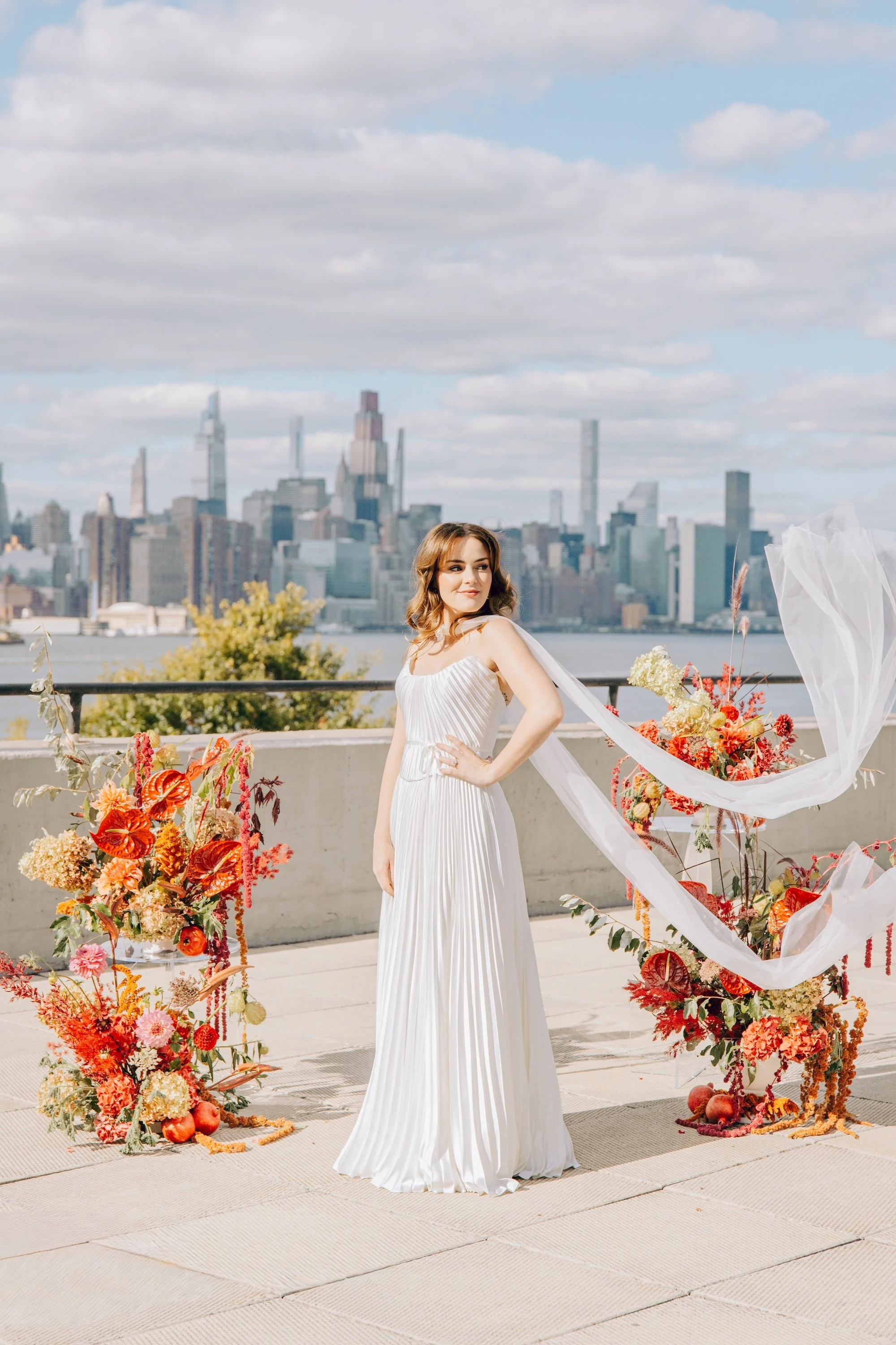 A woman in a white pleated dress stands outdoors on a terrace with a city skyline in the background, surrounded by vibrant fall floral arrangements. marigolds florals nyc florist wedding florals the w loft wedding pedestal wedding ceremony anthurium 