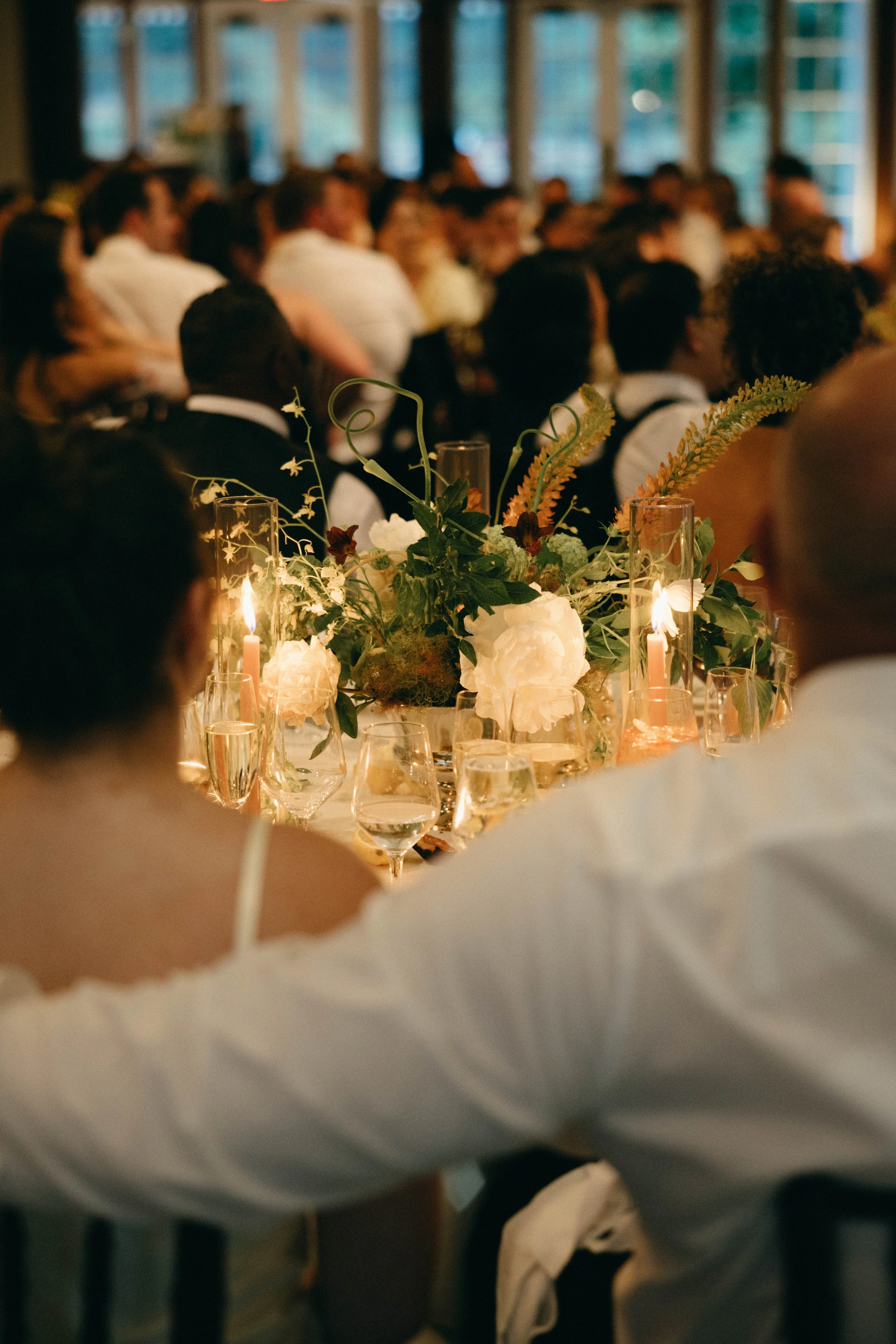 Blurred image of a large group of people attending a formal event or wedding reception, seated at long banquet tables with floral centerpieces and lit candles. marigolds florals nyc florist wedding florals central park boathouse wedding