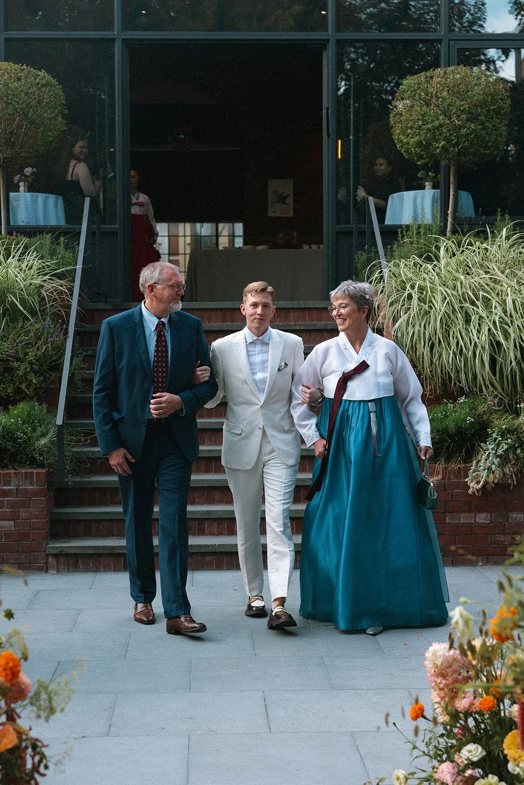A young man in a white suit walking with his parents, who are dressed in formal traditional and business attire, down outdoor steps surrounded by greenery and flowers, with a building and people seen through windows behind them. marigolds florals nyc