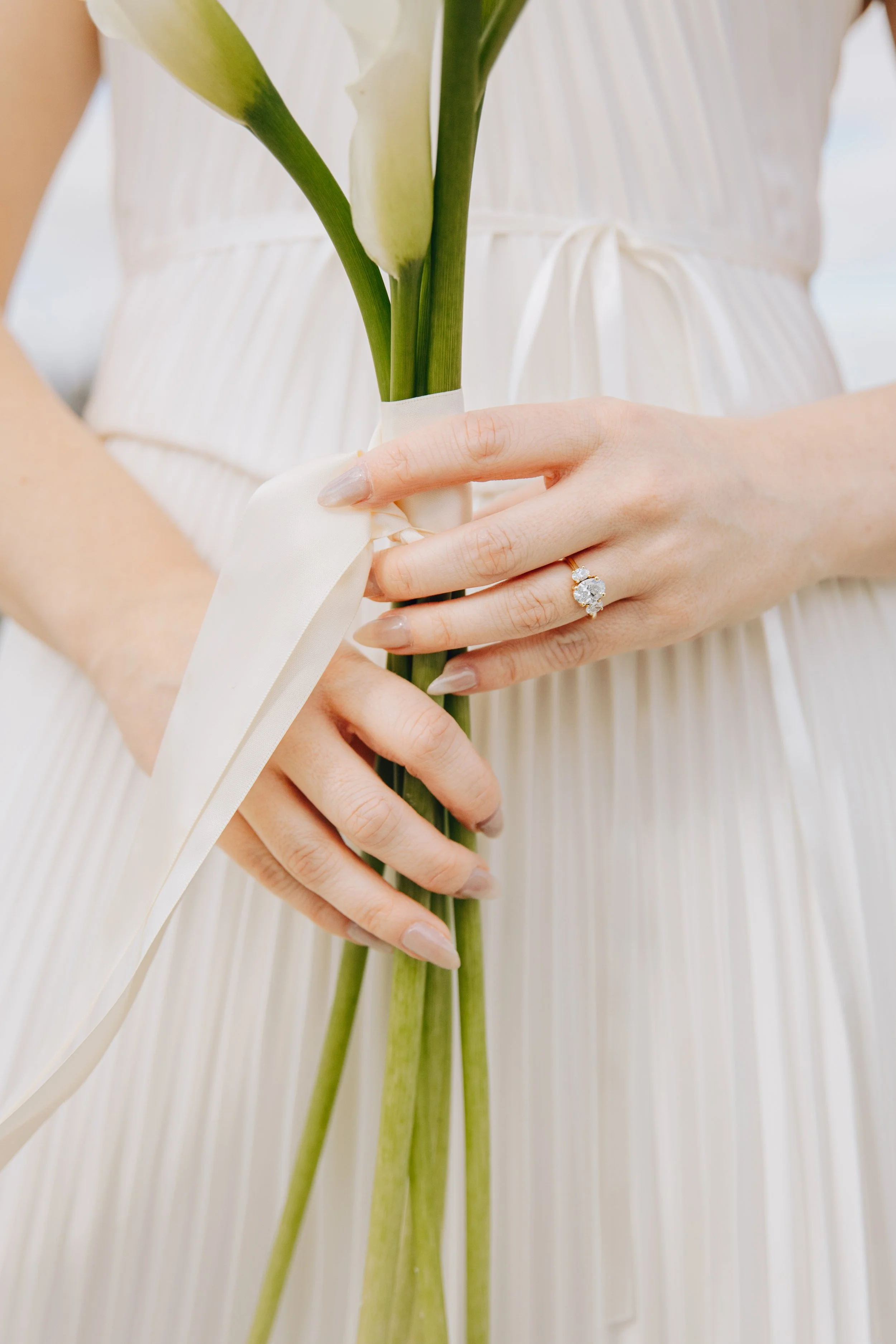 A person holding a bouquet of white calla lilies tied with a white ribbon, wearing a white dress and a ring with a large gemstone.