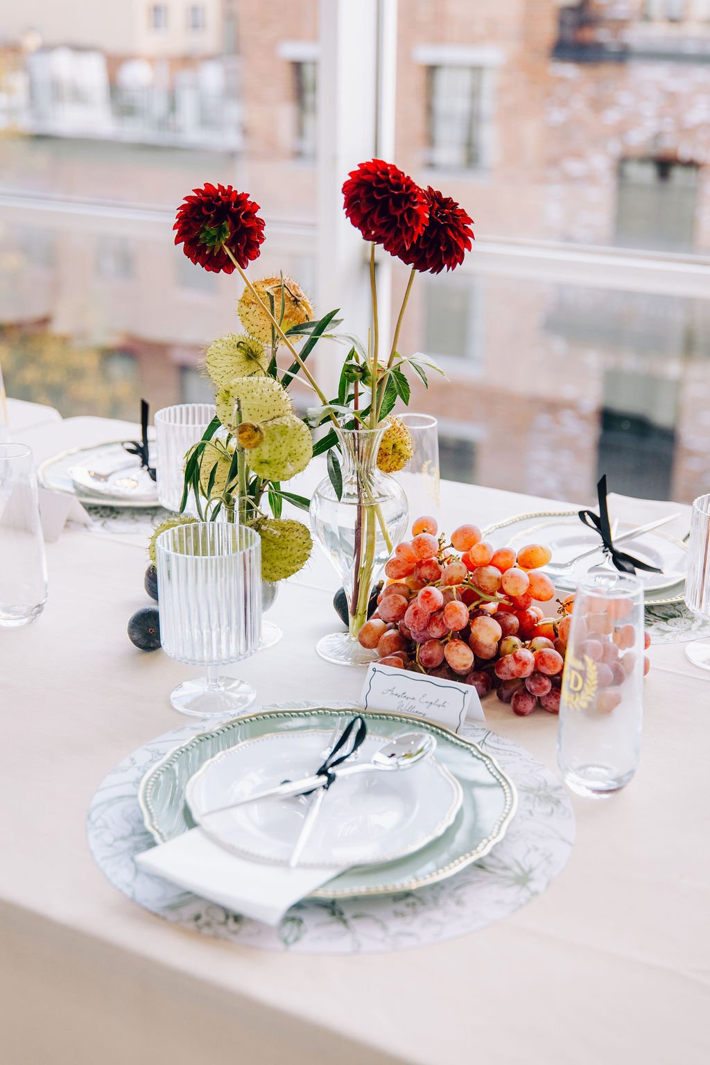 Elegant table setting with white plates, glasses, and cutlery, decorative flowers in a vase, and a bunch of grapes on a light-colored tablecloth in a bright room with large windows and a city view. marigolds florals nyc florist wedding florals 