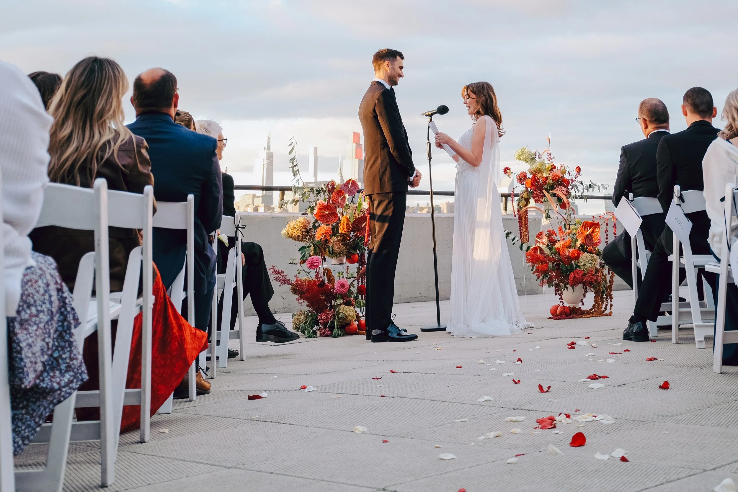 A wedding ceremony with a bride and groom standing at an altar outdoors, surrounded by guests seated on white chairs. The bride is wearing a white dress, and the groom is wearing a brown suit. The background shows a cityscape with tall buildings and 