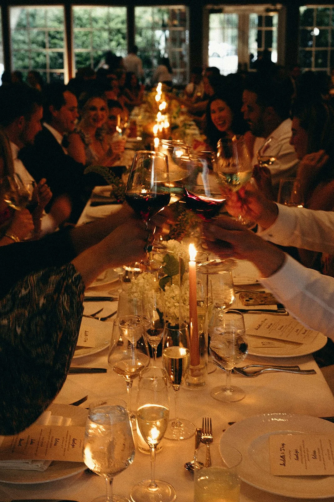 People at a dinner party raising glasses of red wine in a toast, with a candle and floral centerpiece on the table, dimly lit indoor setting. marigolds florals nyc florist wedding florals central park boathouse wedding