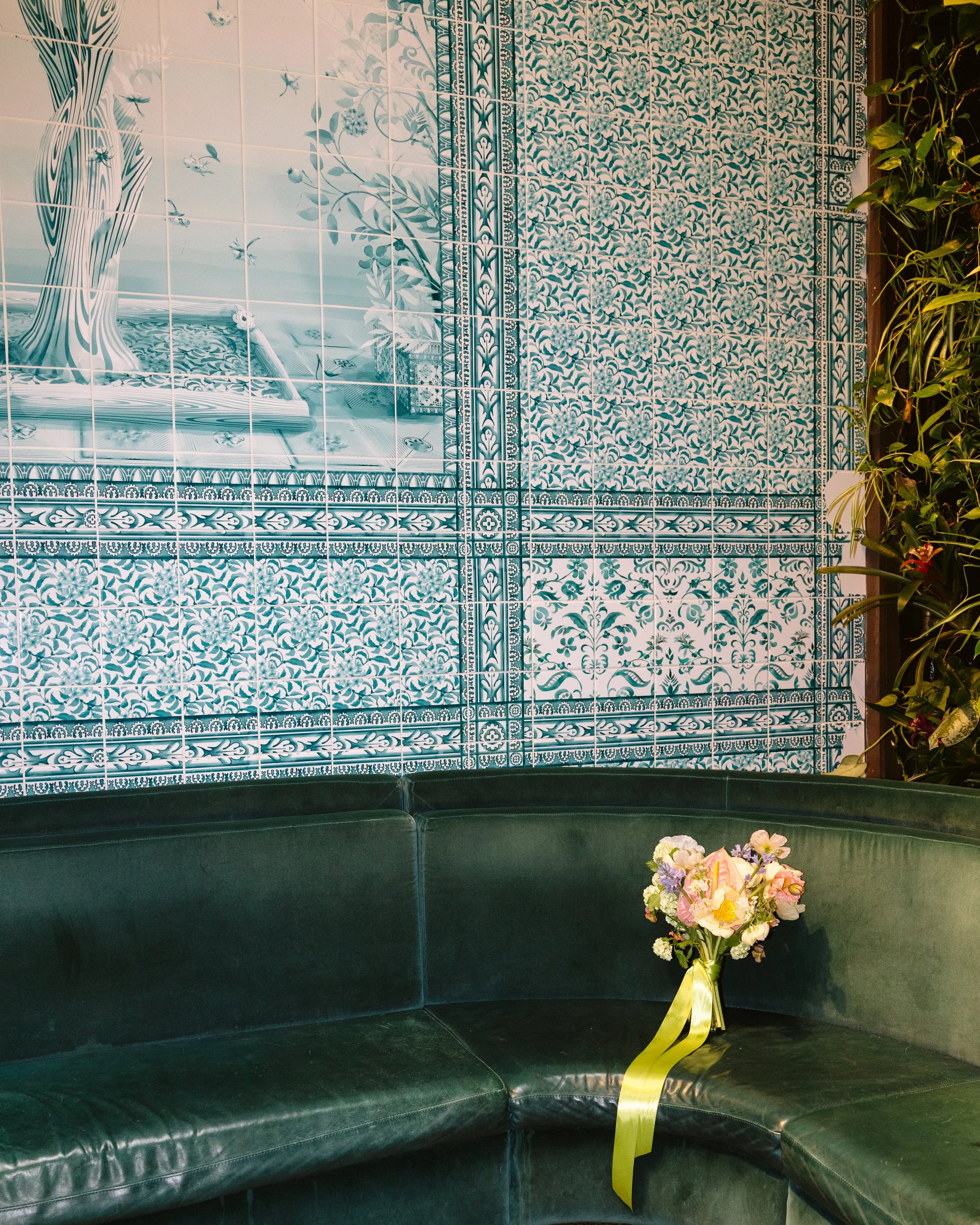A flower bouquet placed on a dark green, semi-circular leather sofa in front of a decorative blue and white patterned tiled wall and some green plants on the side. marigolds florals nyc florist wedding florals houston hall nyc bar wedding
