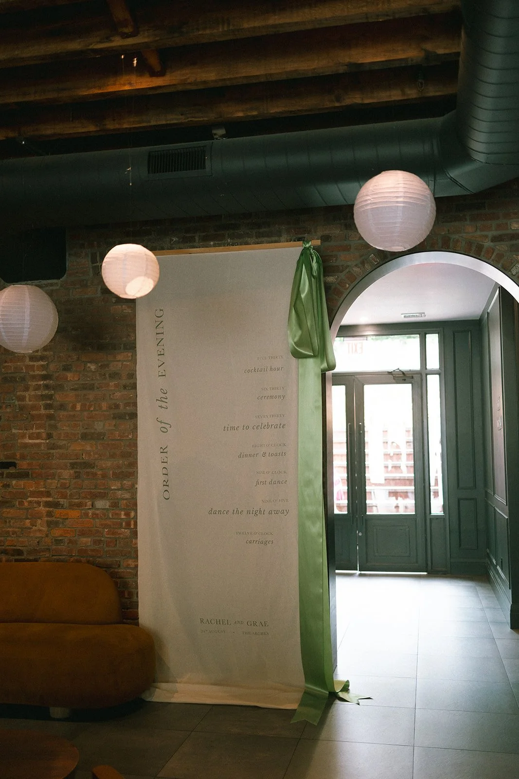 Indoor venue with brick wall, white hanging paper lanterns, green door, and a white banner with a schedule for a wedding or celebration event. marigolds florals