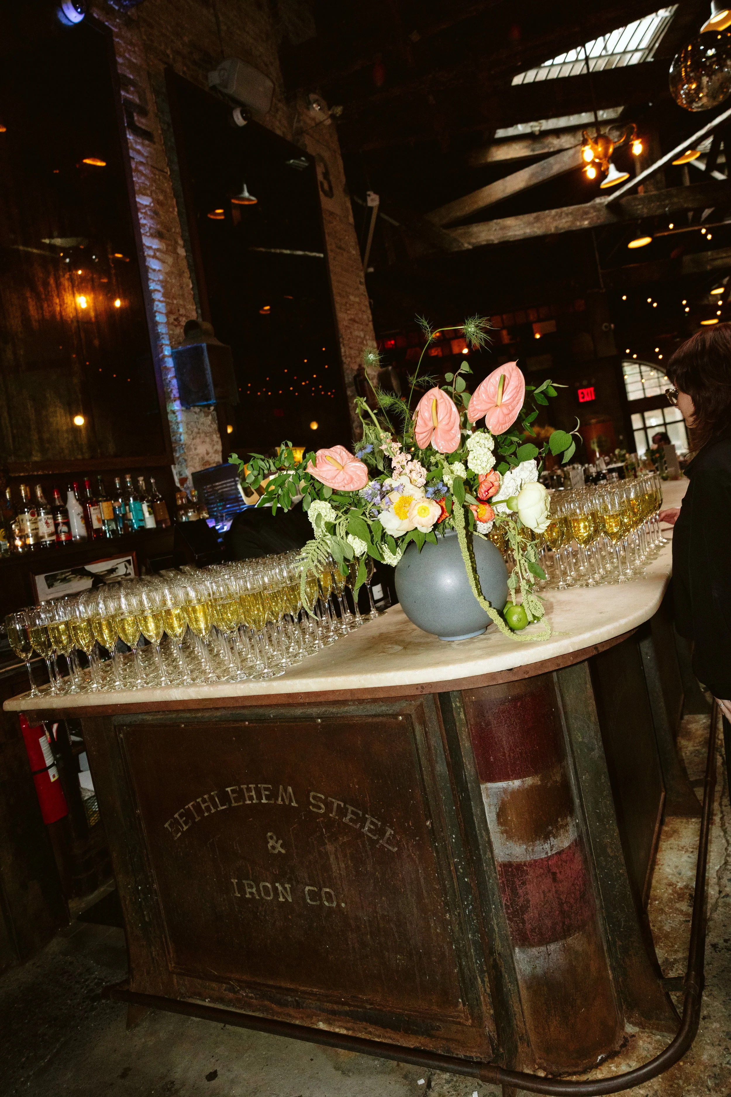 A bar counter with a large floral arrangement in a grey vase, featuring pink anthuriums, white roses, and green foliage, with glasses of champagne or white wine lined up in front. The background shows a rustic interior with exposed brick walls, dark 