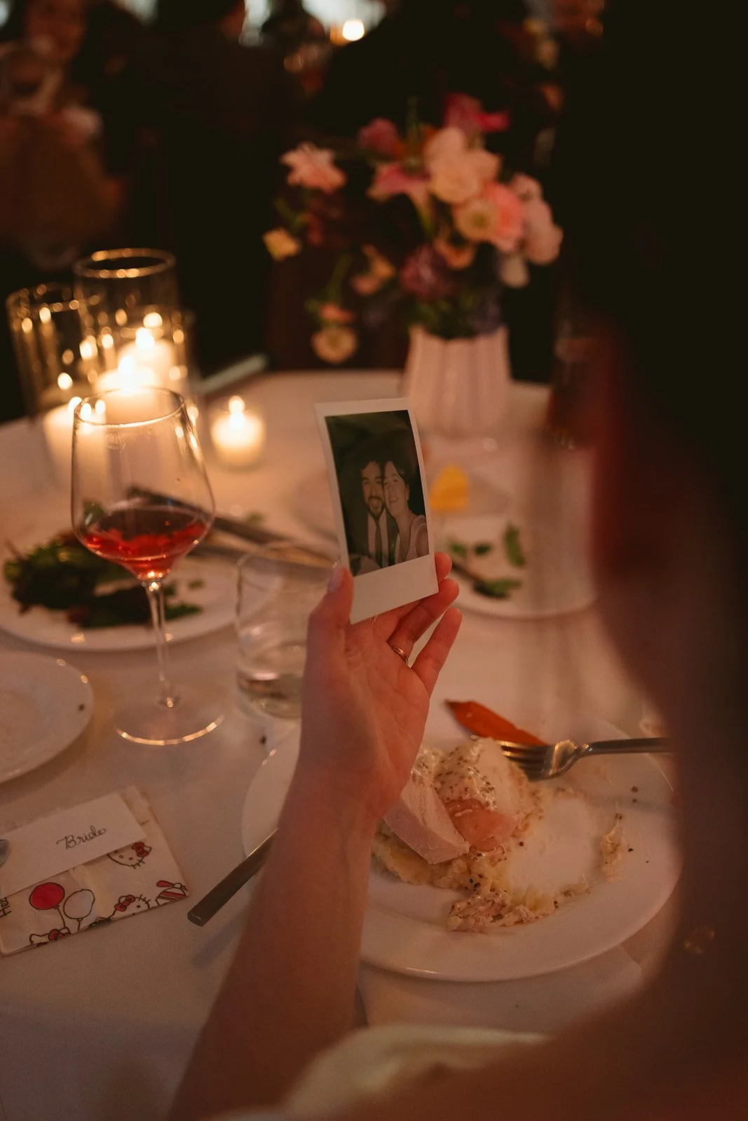 A person holding a Polaroid photo of two smiling people at a dinner party. The scene includes a table with a glass of red wine, a plate with leftover food, a vase of pink and white flowers, lit candles, and a card that says 'Bride'. marigolds florals