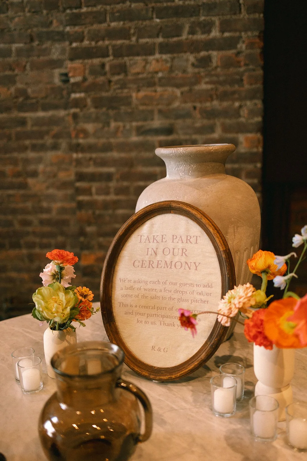 A decorative table setting with a large ceramic vase, small vases with colorful flowers, glass candle holders with white candles, and a framed sign with details about a wedding ceremony, all against a brick wall background. marigolds florals