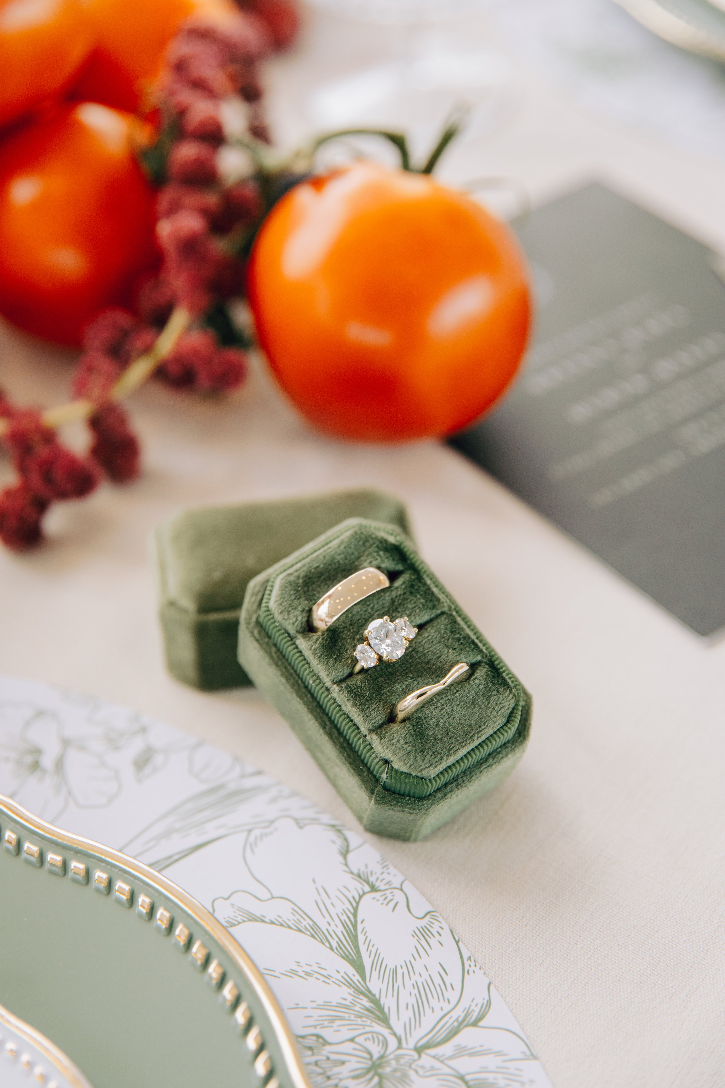 A green velvet ring box with three rings inside, a large sparkling diamond ring, a thin gold band, and a wavy silver ring, placed on a table with floral decorative paper, surrounded by a gray plate and some fruits including a tomato, a bunch of grape