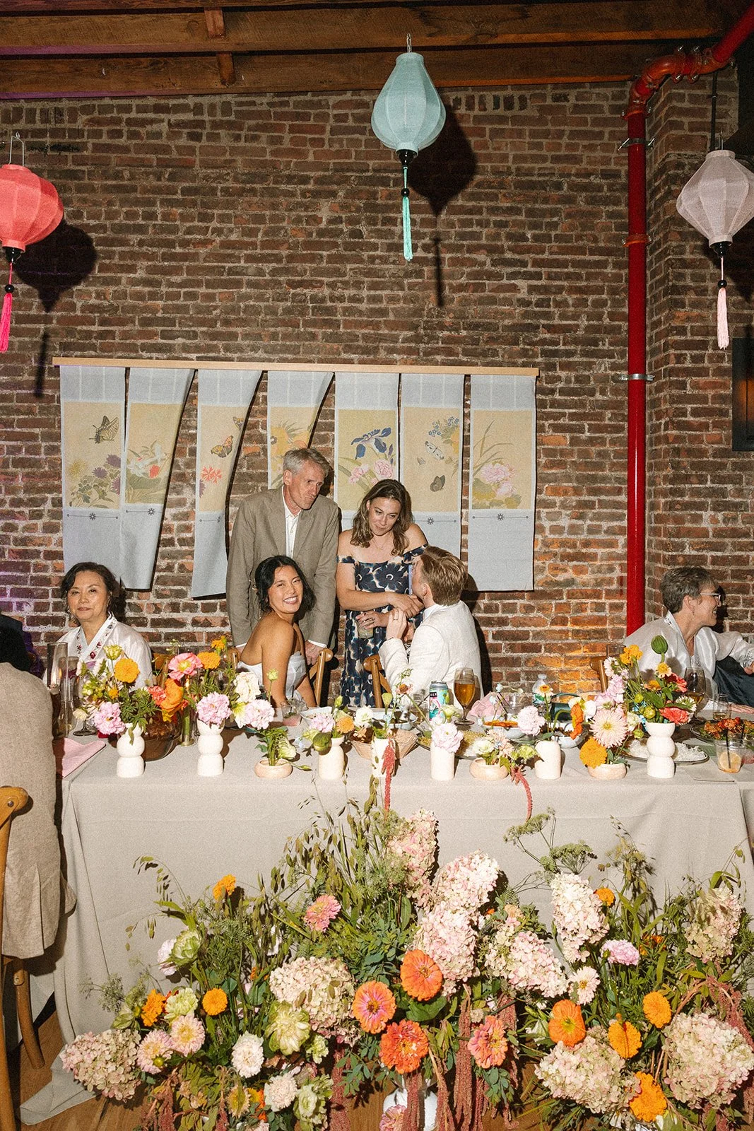 People celebrating at a wedding reception, with a decorated table and floral arrangements, against a brick wall with lanterns hanging from the ceiling. marigolds florals nyc florist wedding florals