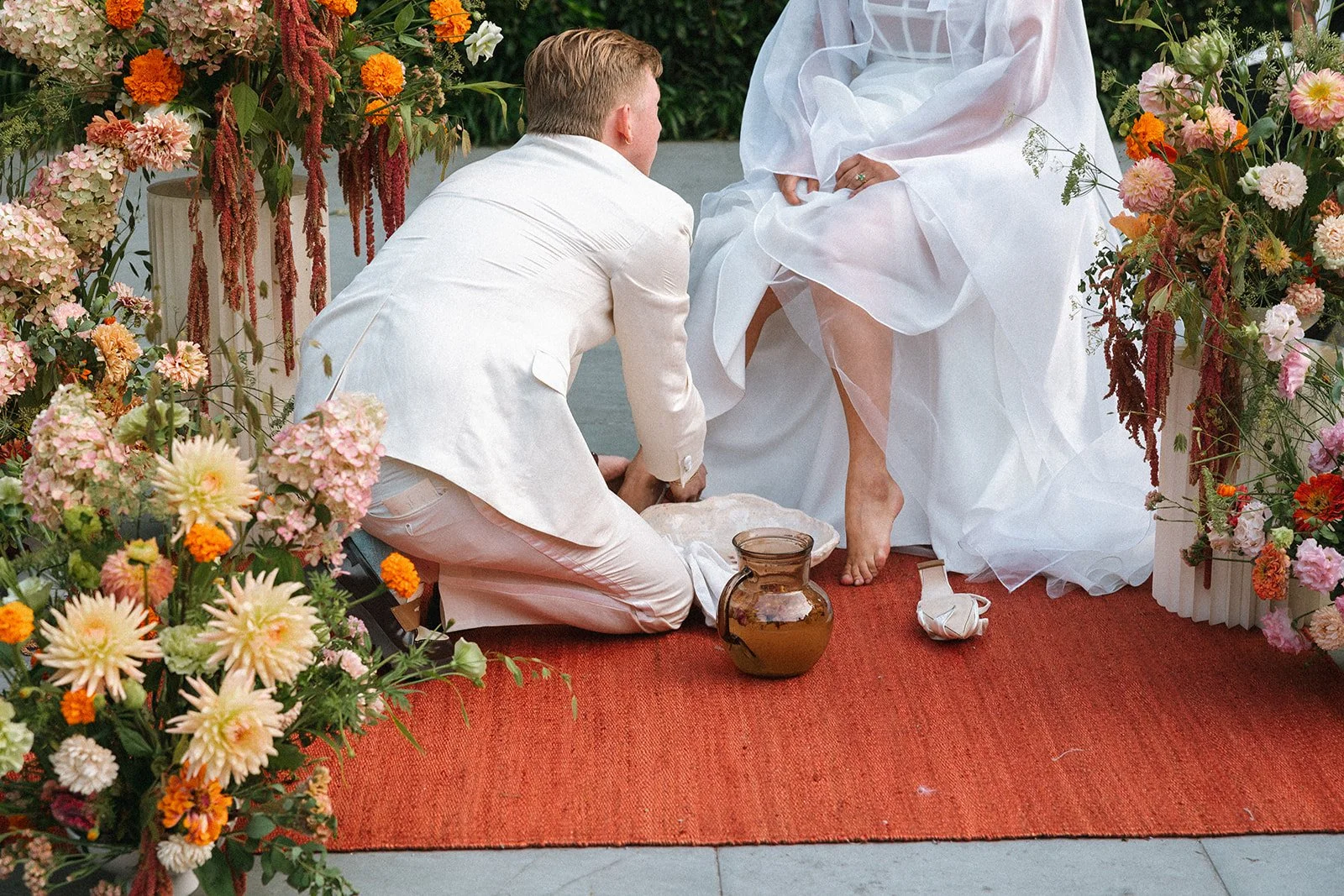 A wedding ceremony with a man in white kneeling on a red carpet, holding a woman's foot, surrounded by colorful flowers. marigolds florals nyc florist wedding florals, dahlias, warn colors, autumn wedding