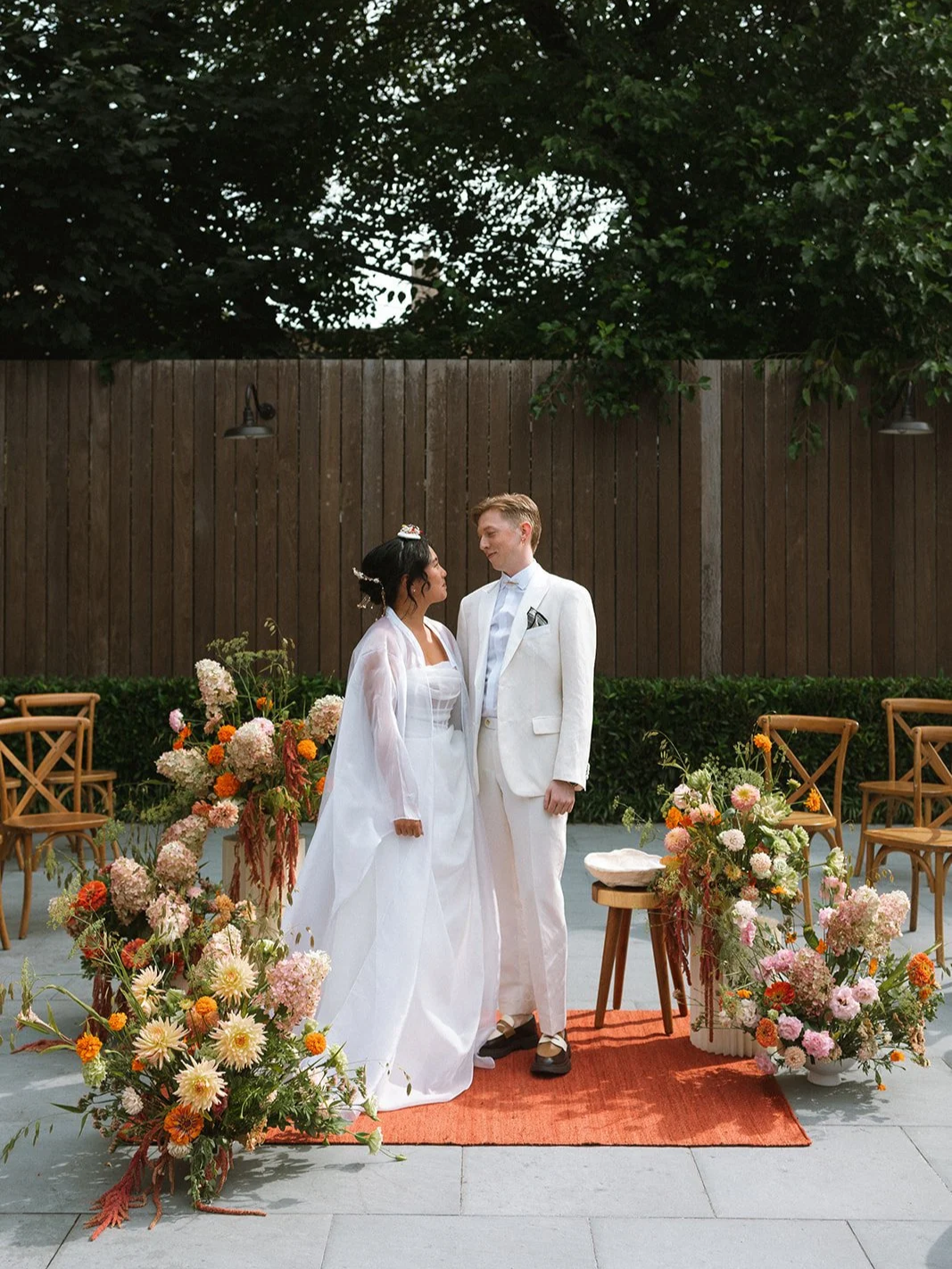 A bride and groom or lgbtqia+ couple standing on an outdoor wedding stage decorated with colorful flowers, facing each other, with chairs and a wooden fence in the background. Wedding at The Arches in Brooklyn, New York
