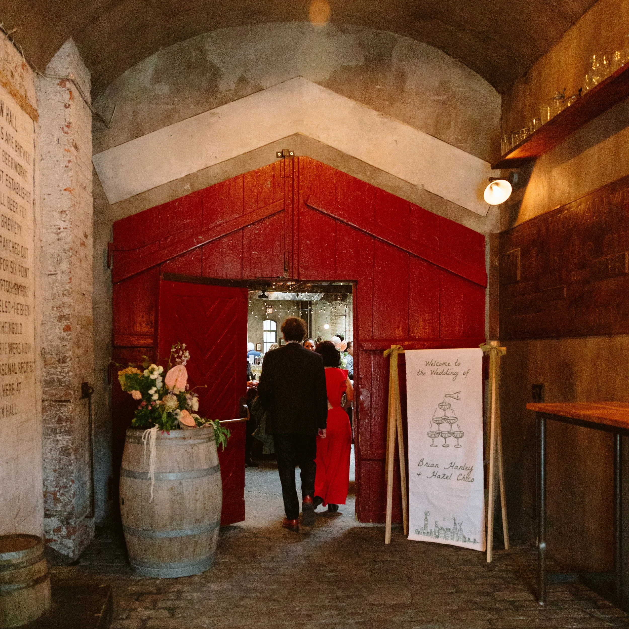 A red wooden archway leading to a wedding reception inside a rustic venue with brick and stone walls. Decor includes a white sign with wedding details and a flower arrangement marigolds florals nyc florist wedding florals houston hall nyc bar wedding