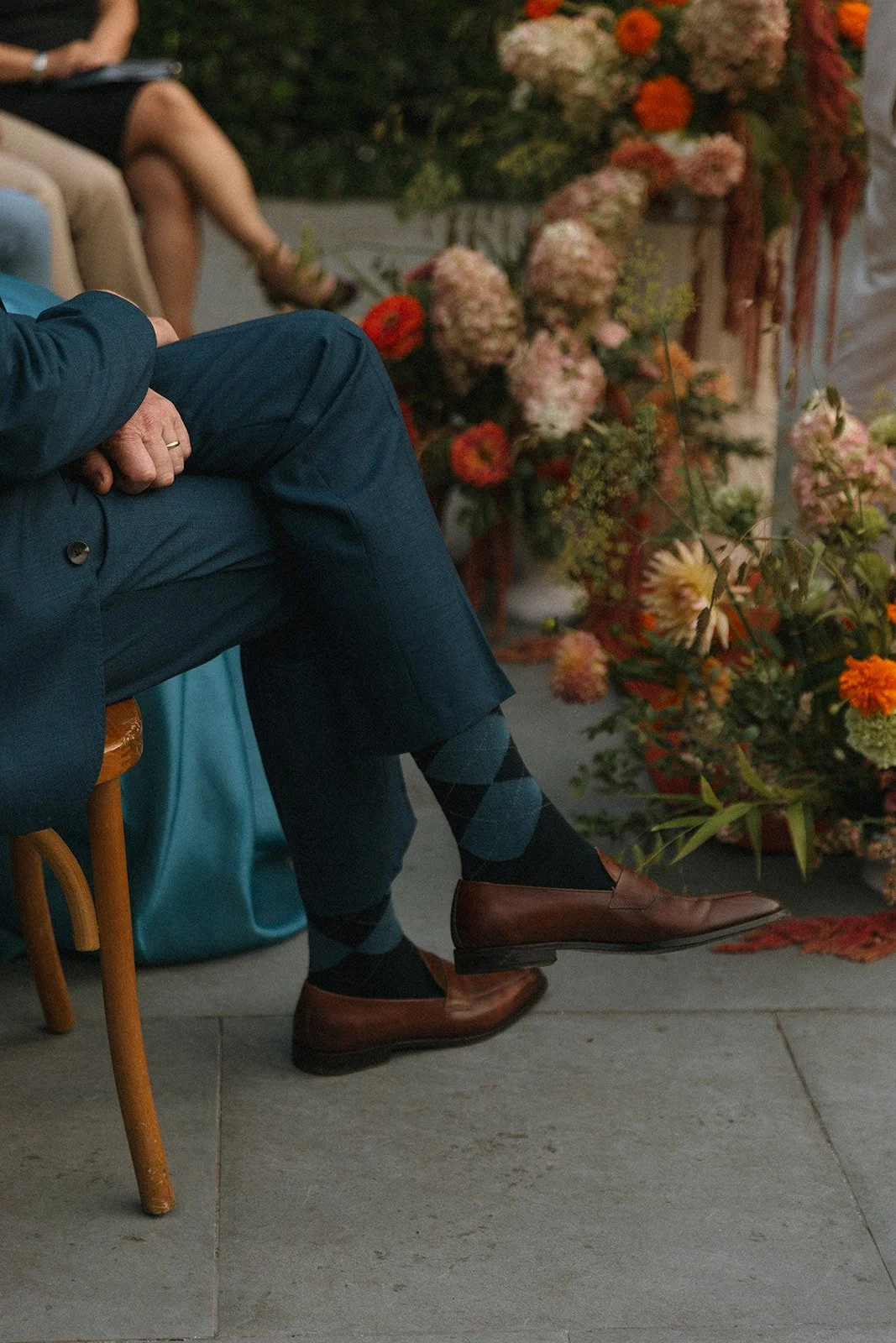 A man in a dark suit with brown dress shoes and argyle socks sitting on a wooden chair at a formal event with a table with flowers in the background. marigolds florals nyc florist wedding florals