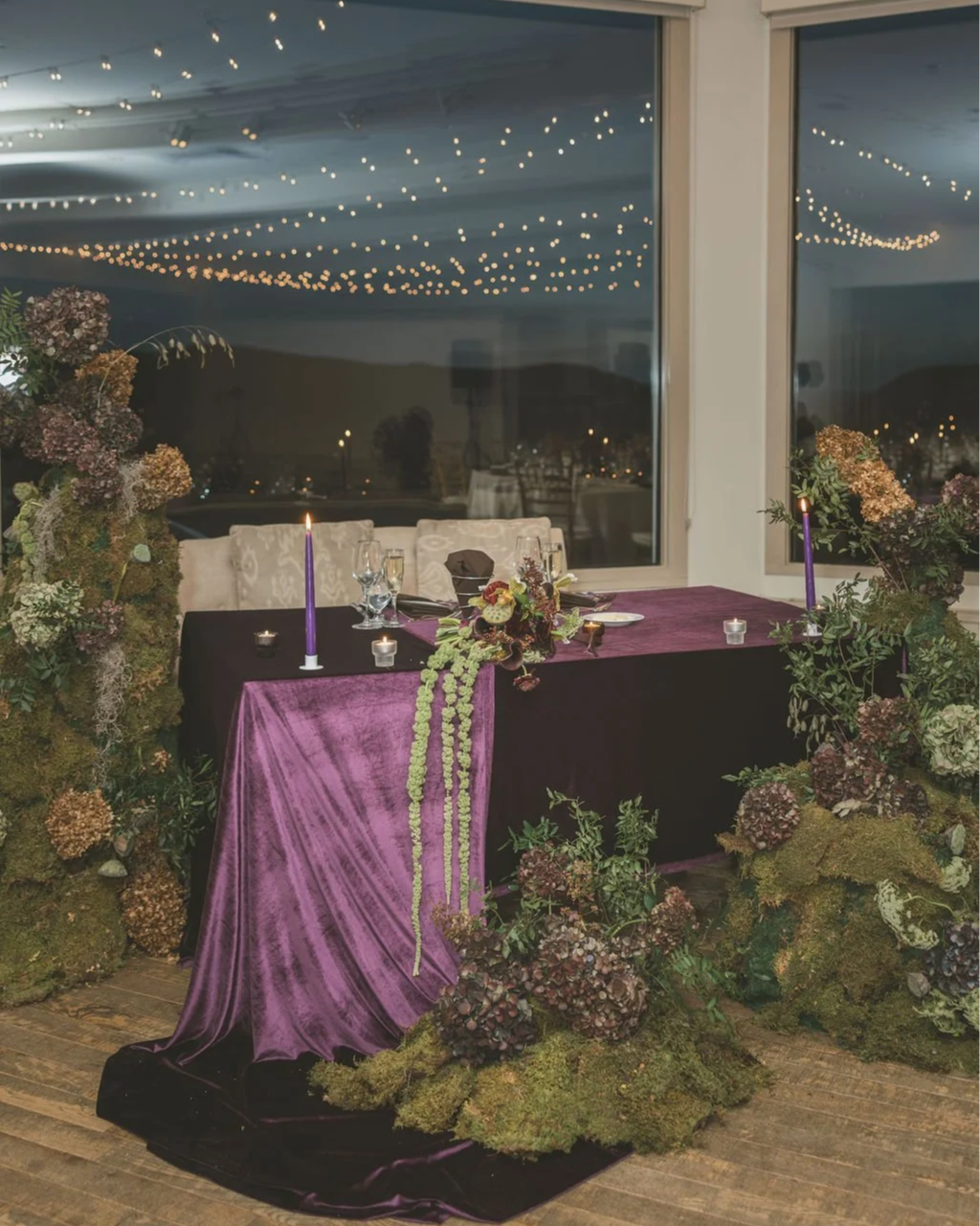 Decorated dinner table with a purple velvet tablecloth, floral centerpiece, candles, and surrounded by moss and hydrangea arrangements, with string lights reflected in the large window behind.