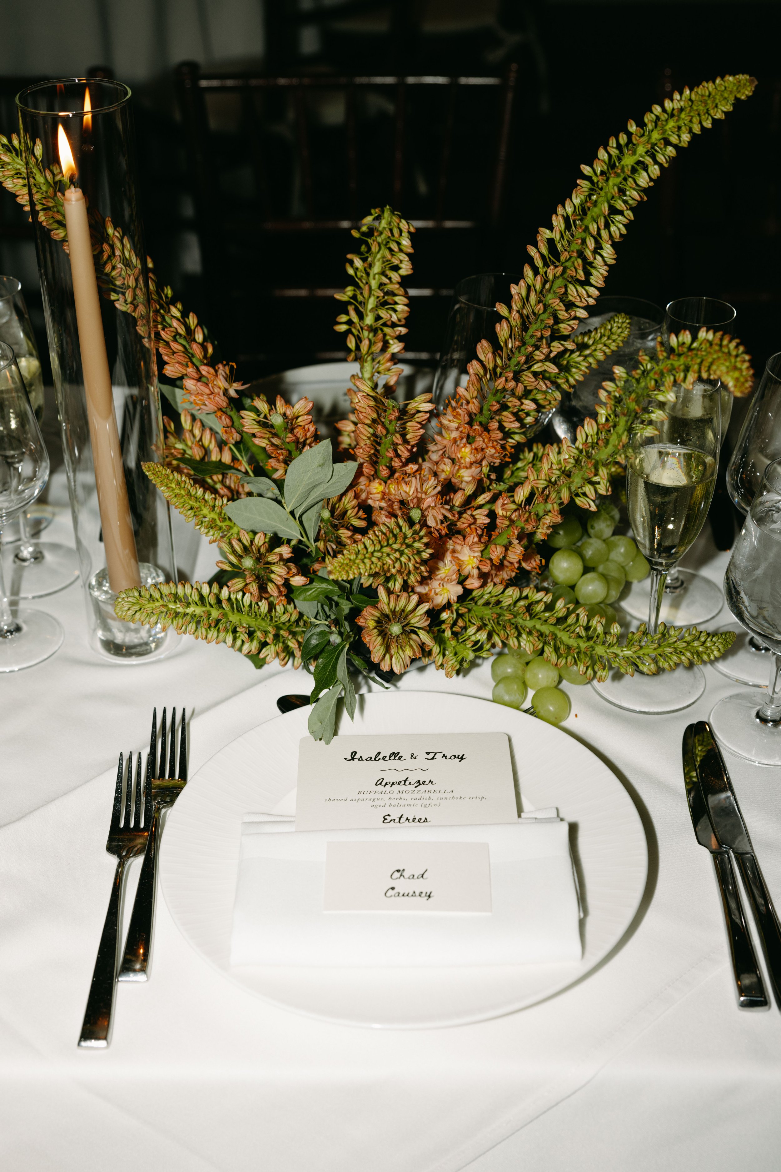 Table setting with a white plate, silverware, a menu card and a floral centerpiece with green grapes, tall candles, and greenery, at an elegant event. marigolds florals nyc florist wedding florals central park boathouse wedding