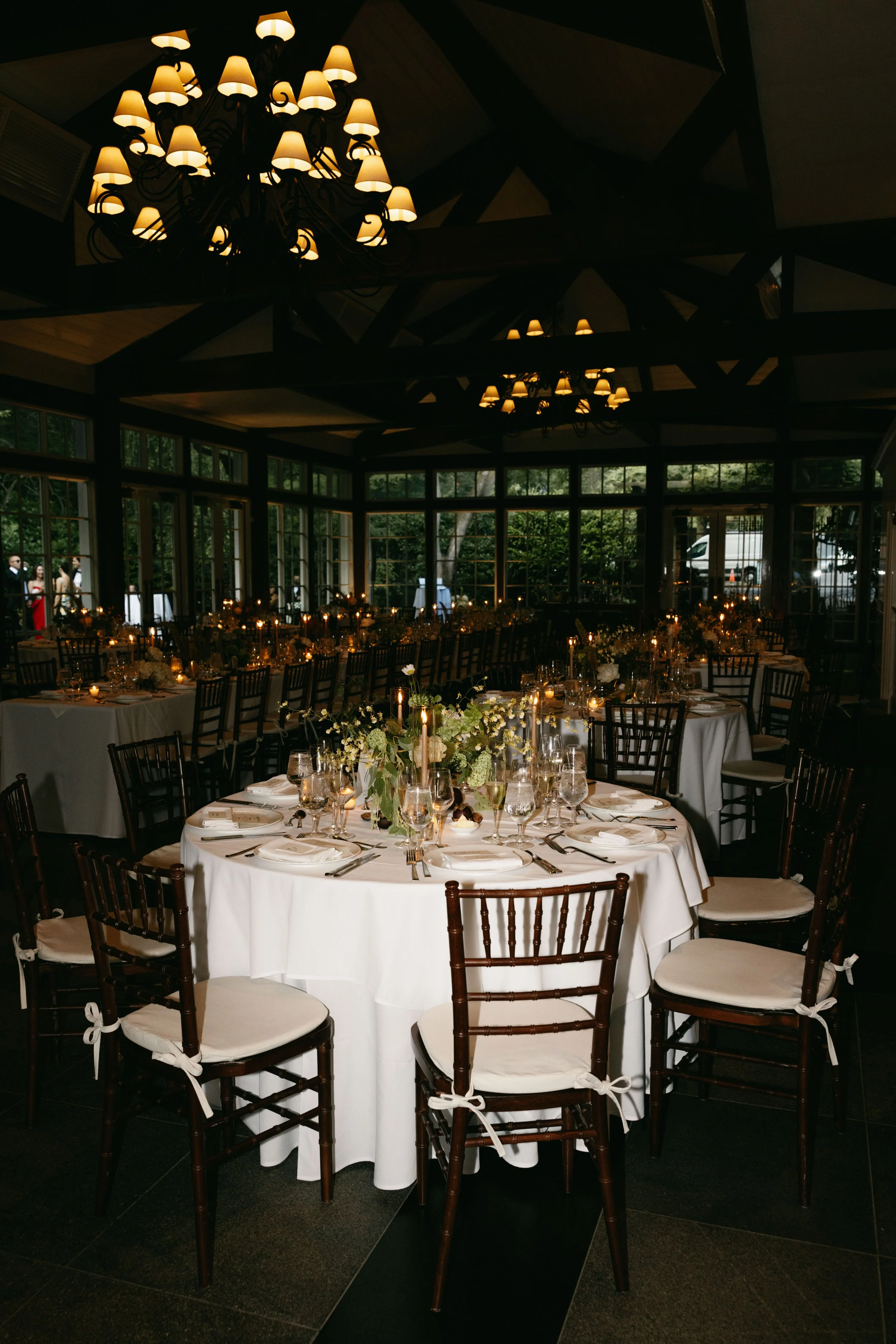 Elegant indoor dining setup with round table draped in a white tablecloth, set with glassware, plates, silverware, and floral centerpiece, surrounded by dark wooden chairs with white cushions, under warm lighting from chandeliers, in front of large w