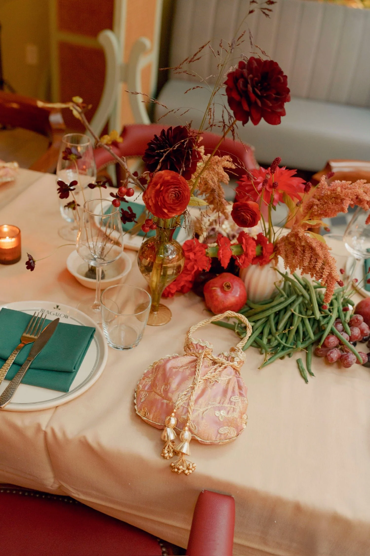 A dining table with floral centerpiece, pink tablecloth, and place settings, including green napkins, wine glasses, and plates. A pink silk pouch with pearl embellishments is also on the table.