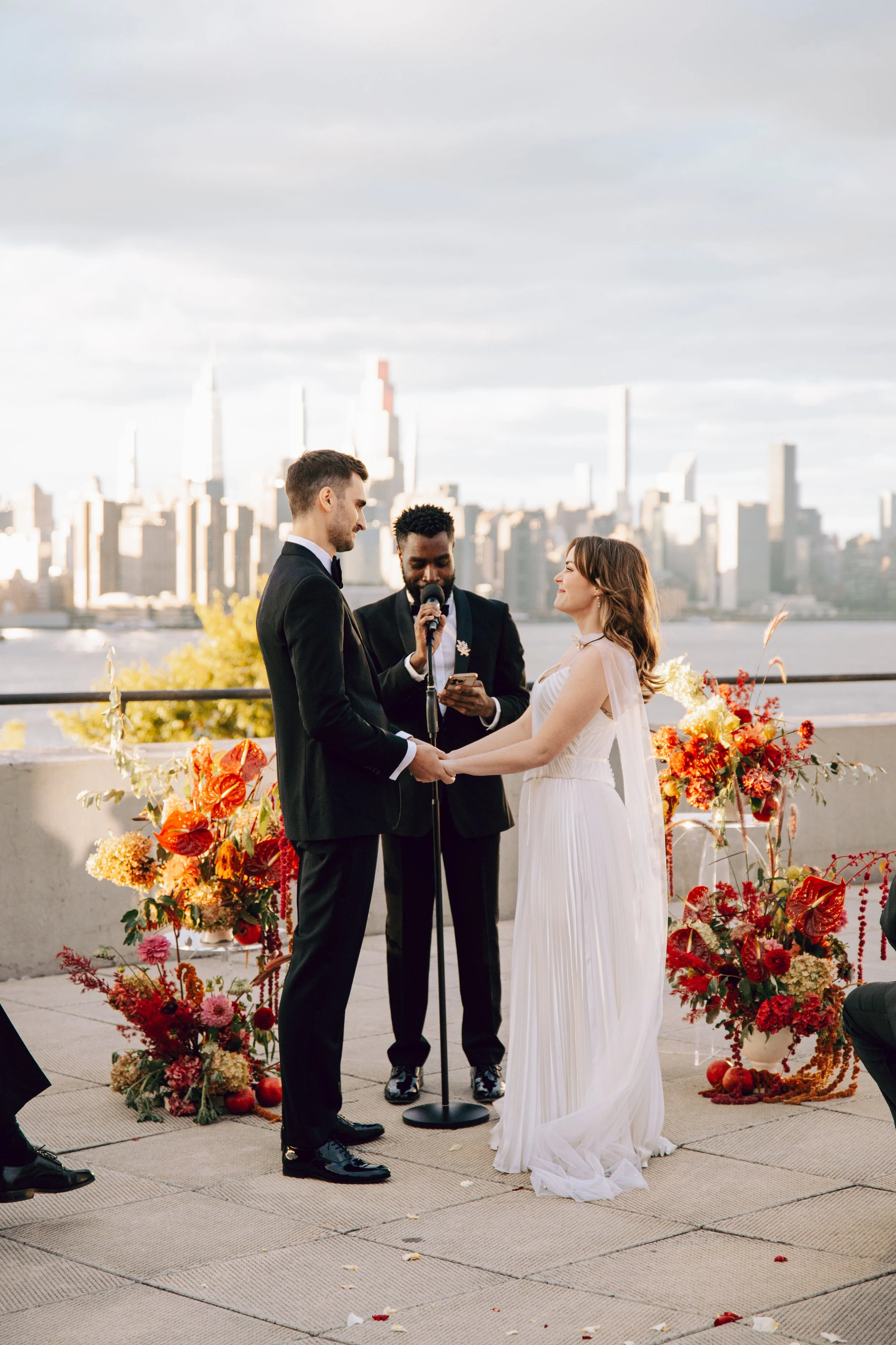 A wedding ceremony taking place outdoors with a city skyline in the background. A bride and groom are holding hands and facing each other, with an officiant reading from a device. The ceremony is decorated with vibrant orange, red, and pink flowers, 