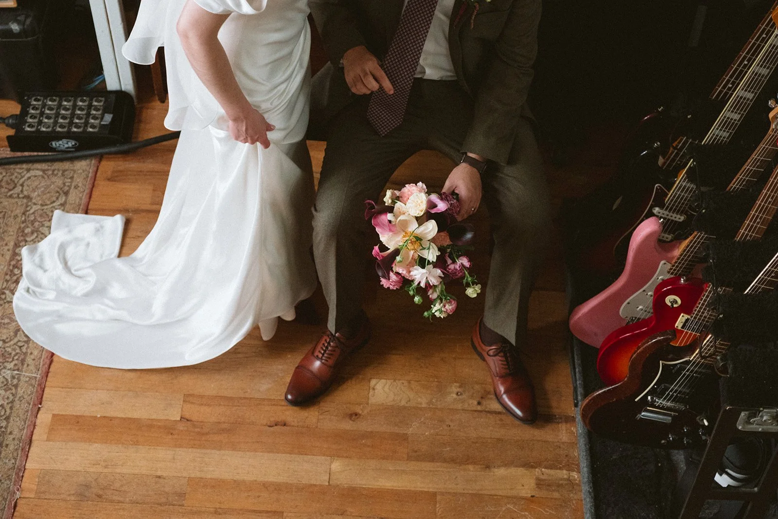 A bride and a man sitting on a bench, holding a bouquet of pink and white flowers. The bride wears a white wedding dress with a train, and the man wears a dark suit with brown shoes. To the right, there are several guitars in a rack.