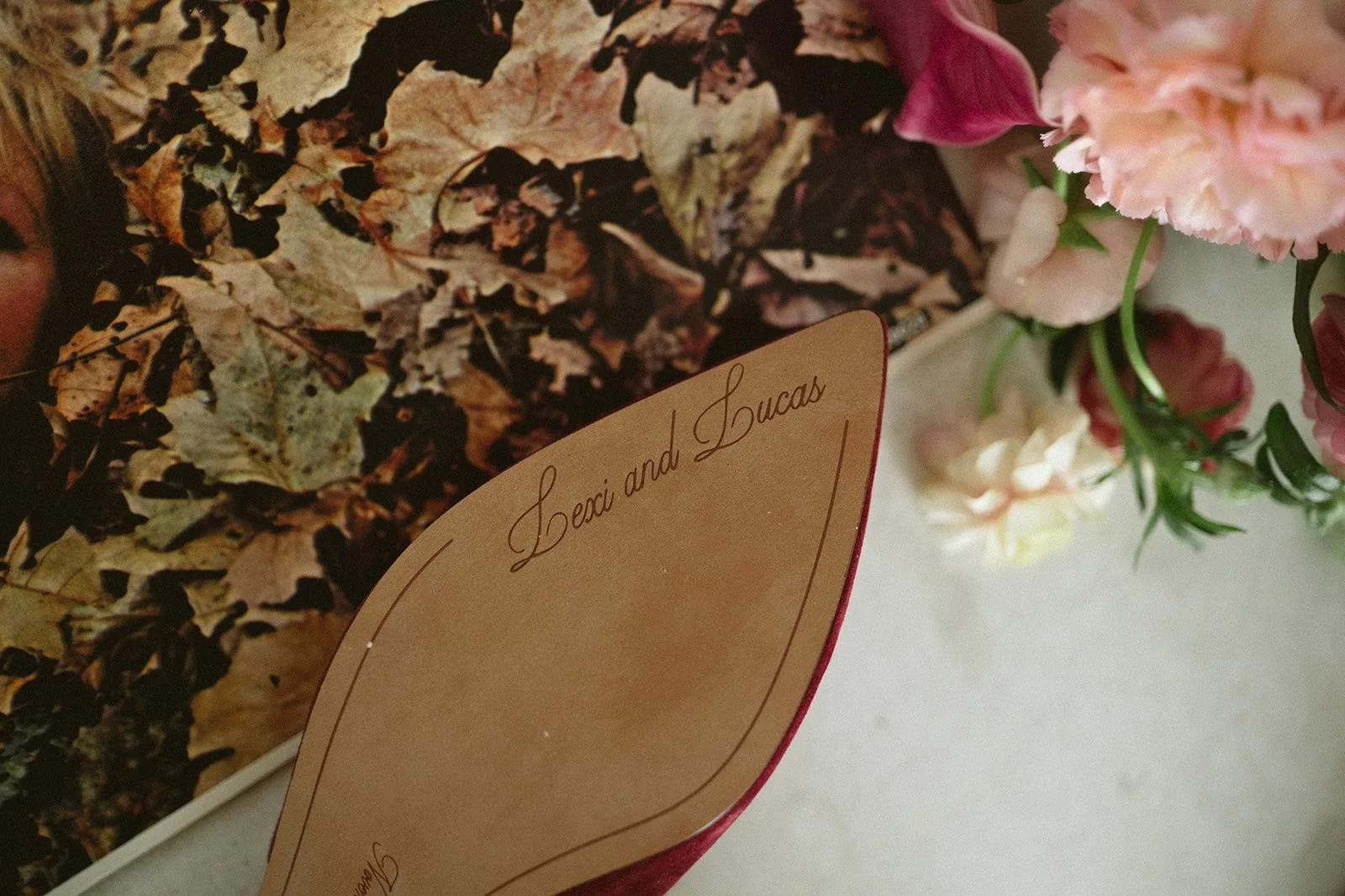 A close-up of a bouquet of flowers with a label that says 'Lexi and Lucas', featuring pink and white flowers with green stems. In the background, there are dried leaves with a mixture of brown, black, and beige colors.