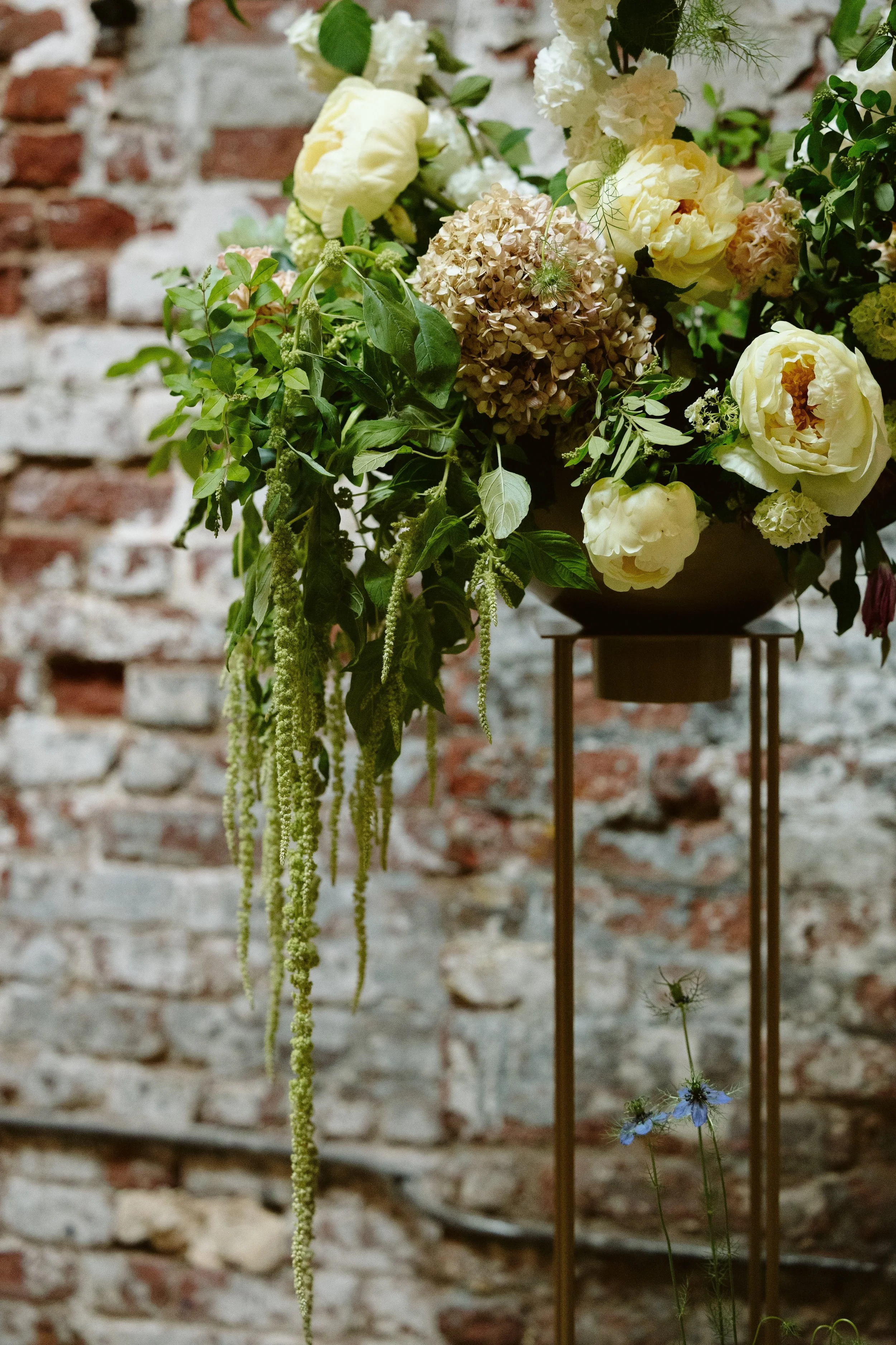A floral arrangement with white, beige, and blue flowers and green foliage in front of a red brick wall. marigolds florals nyc florist wedding florals houston hall nyc bar wedding