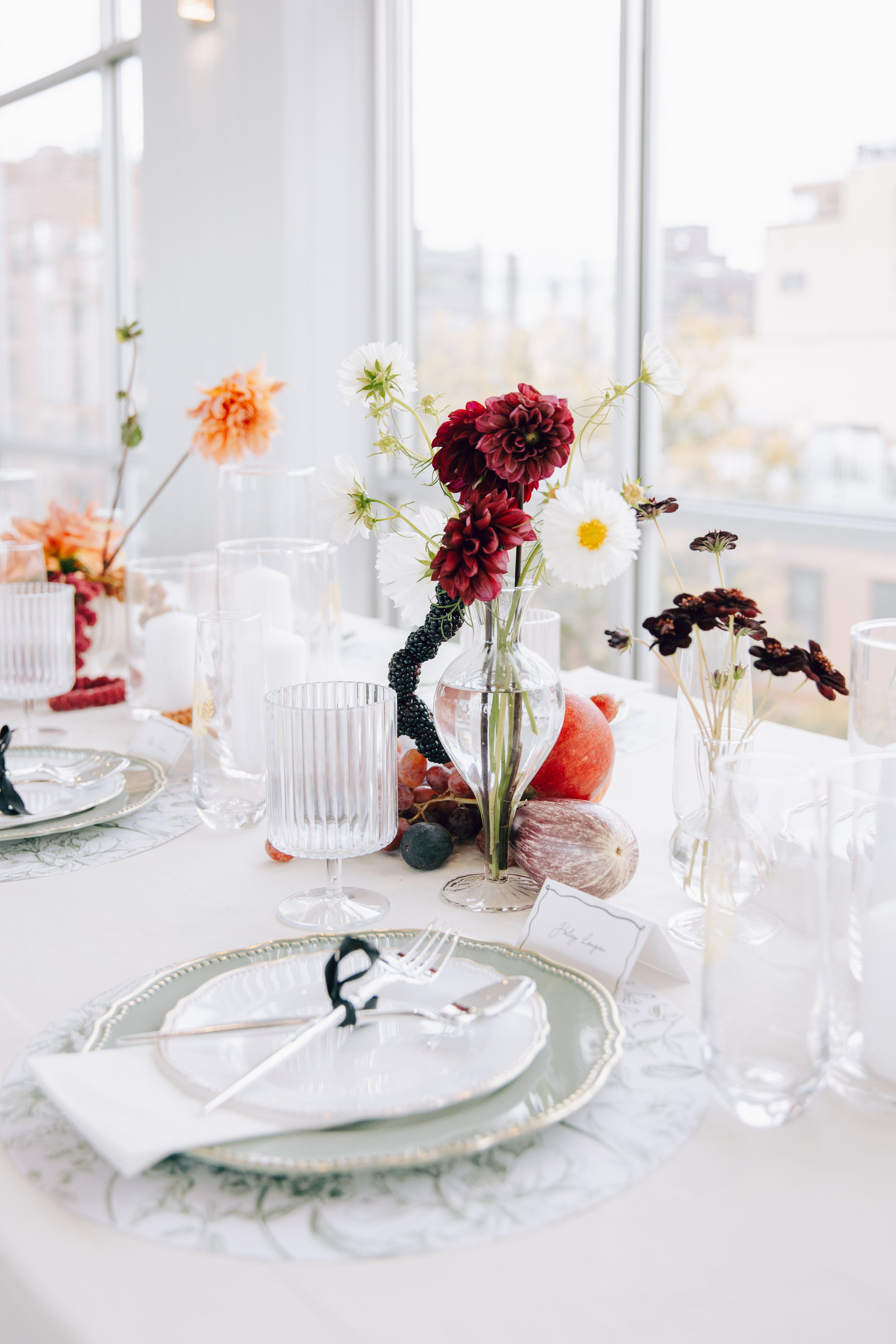 Elegant table setting with a white tablecloth, glassware, white dishware with gold trim, and a floral centerpiece of white, red, and dark purple flowers in a clear vase. There are also some fruits and a name card on the table.