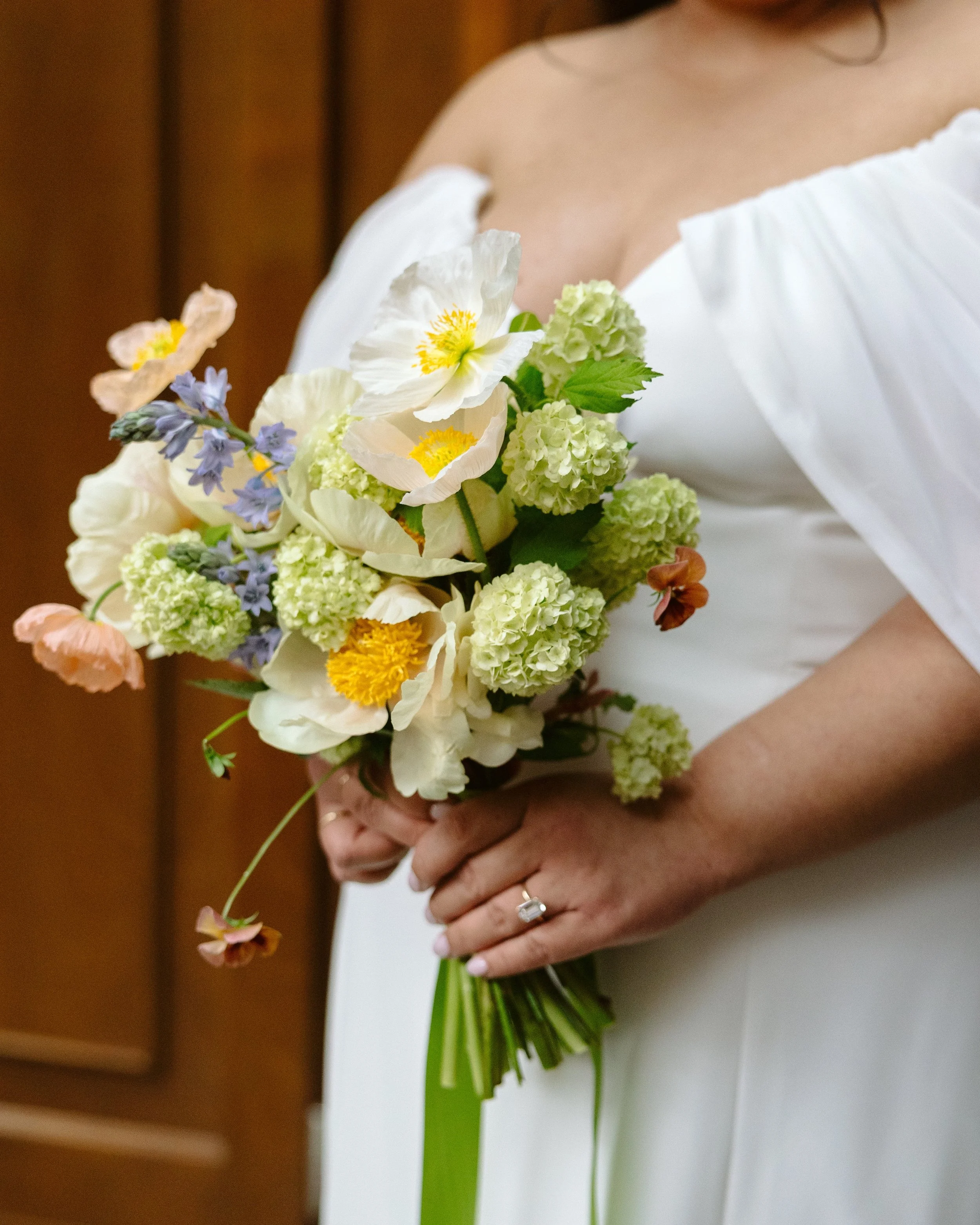 A person in a white off-shoulder dress holding a colorful bouquet of flowers with a wedding ring on their left hand.