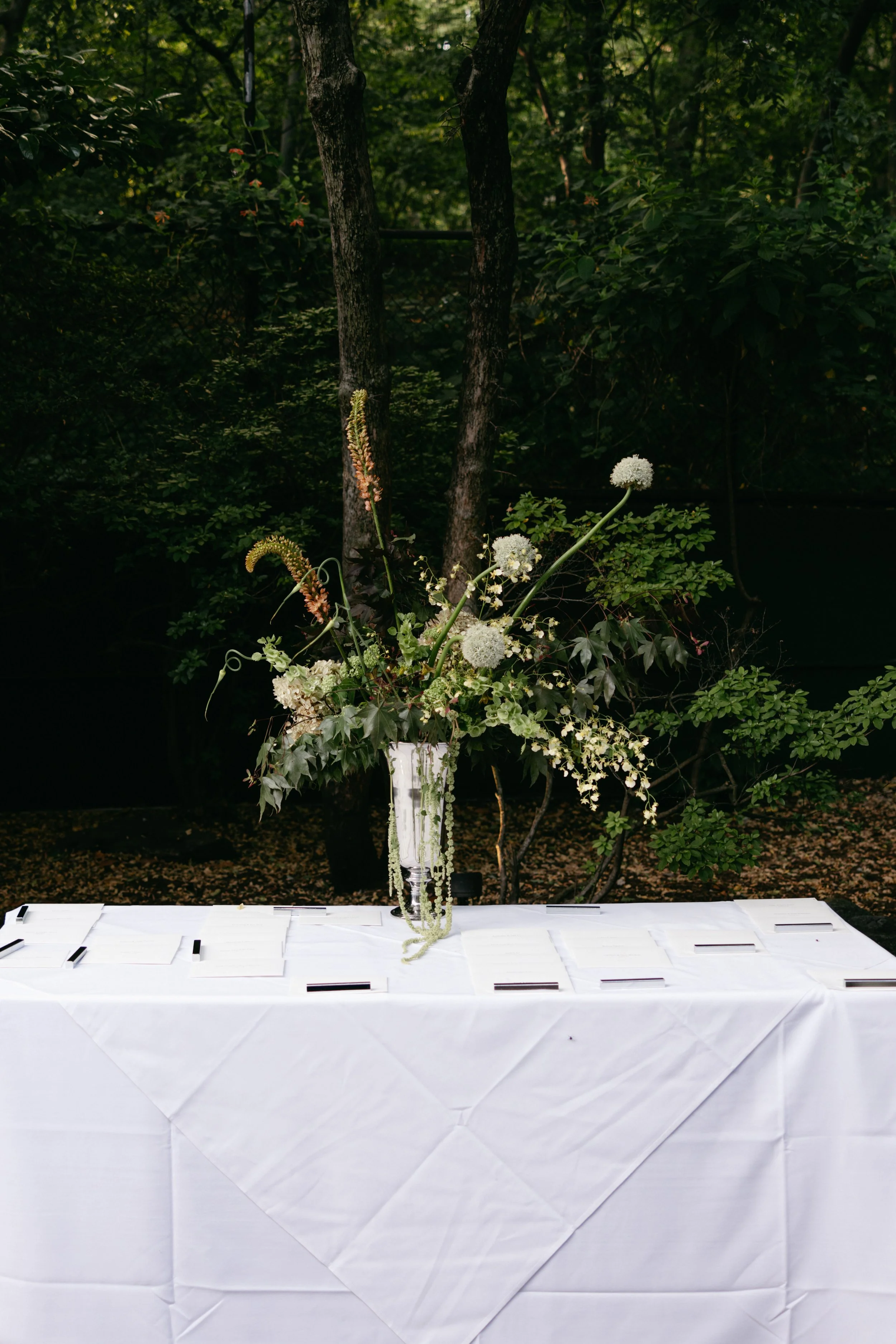 A white table with multiple small envelopes and a tall transparent vase filled with white and green floral arrangements, set outdoors with lush green trees in the background. marigolds florals nyc florist wedding florals central park boathouse