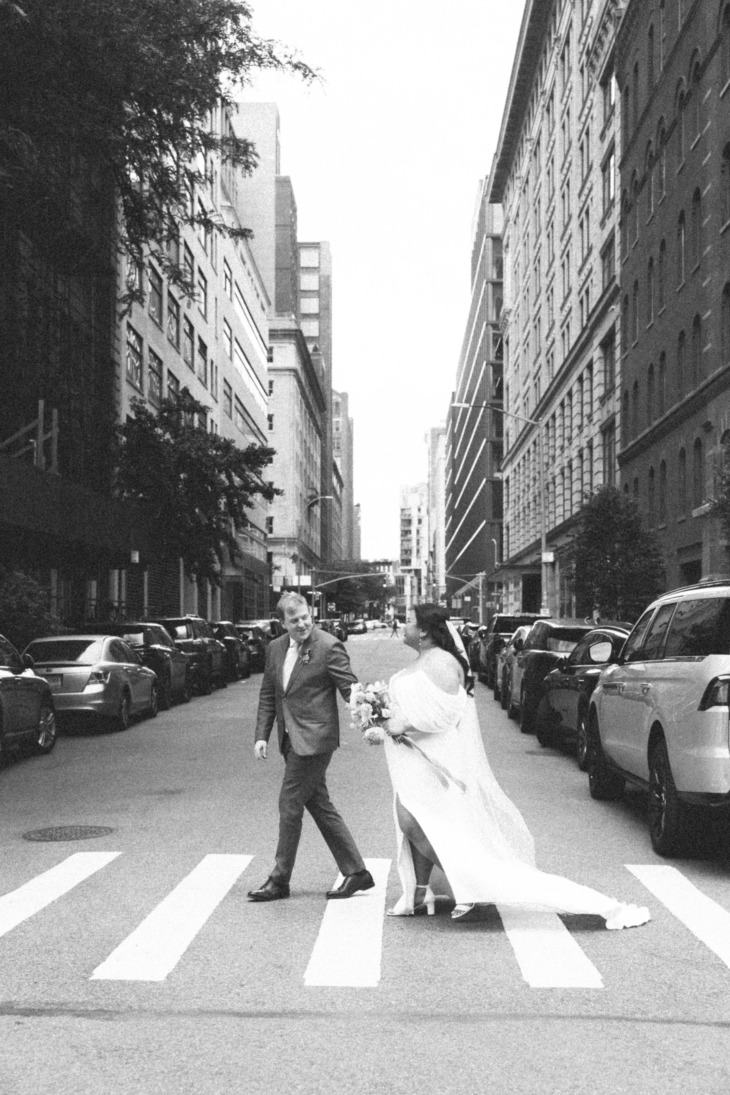 A black-and-white photo of a bride and groom crossing a city street on a crosswalk. The groom is wearing a suit, and the bride is in a wedding gown holding a bouquet. Tall buildings line the street, with parked cars on both sides and trees along the 