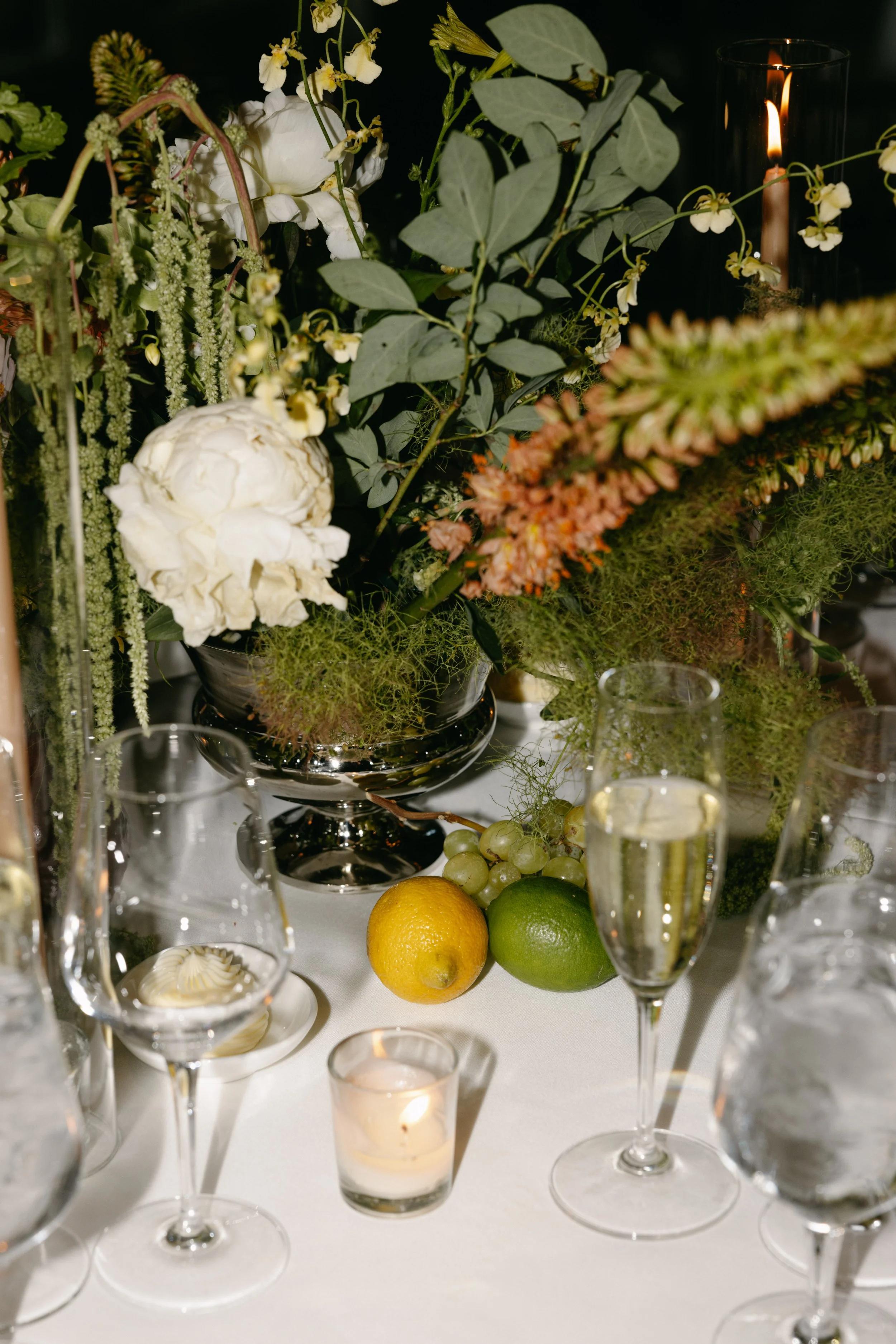 Elegant table centerpiece with white and blush flowers, greenery, and candles surrounded by champagne flutes and candles on a white tablecloth. marigolds florals nyc florist wedding florals central park boathouse wedding