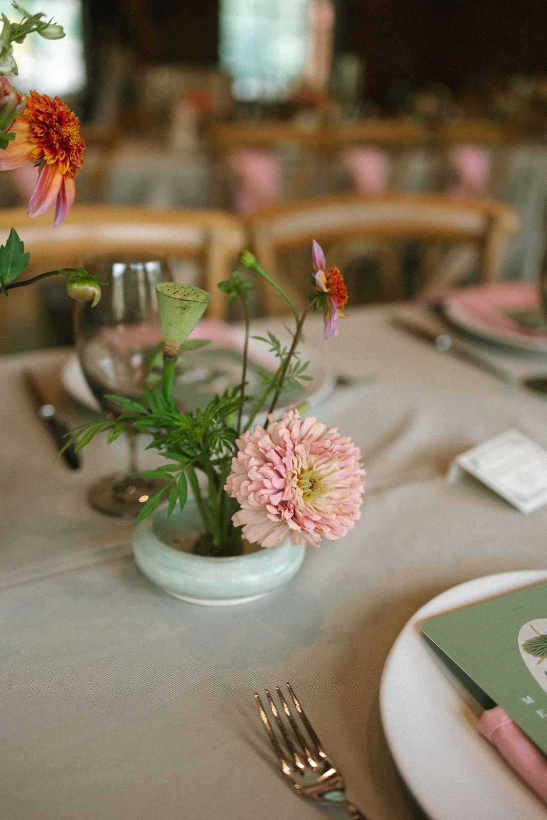 A table centerpiece with pink flowers in a white vase, set on a beige tablecloth at a dining table with gold utensils and layered plates in a softly lit restaurant or event space. marigolds florals
