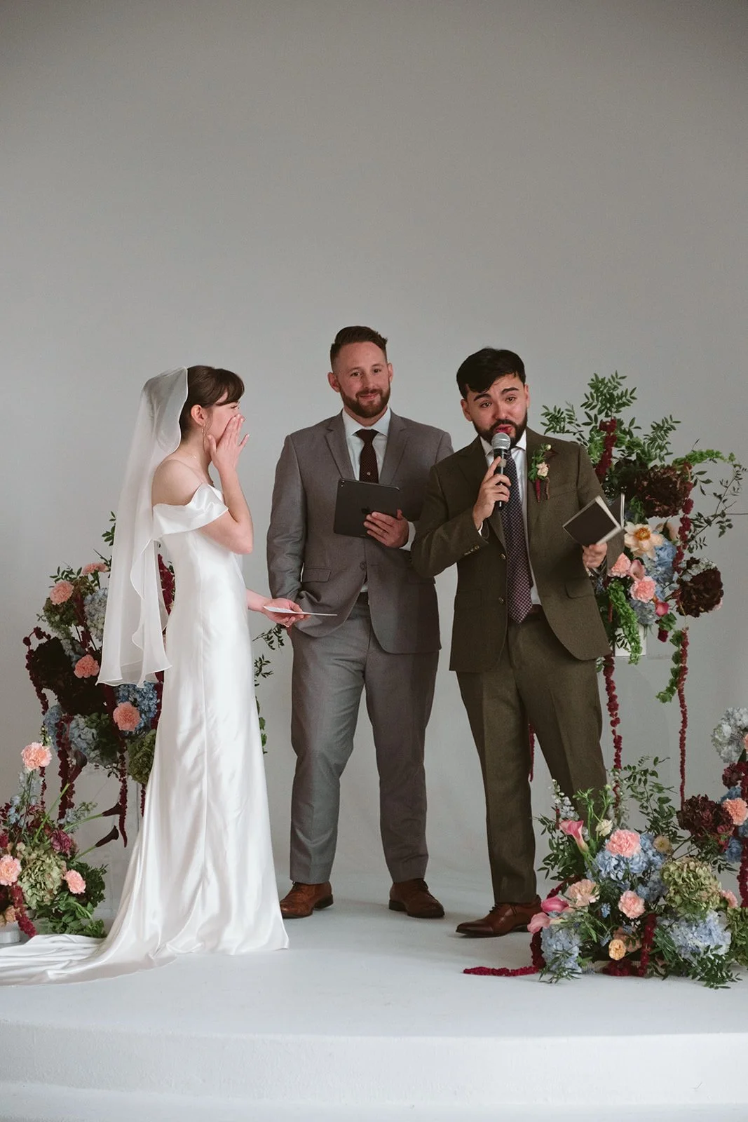 A wedding ceremony with a bride in a white dress and veil, and two men in suits, one with a microphone, standing in front of floral arrangements. marigolds florals nyc florist wedding florals atmosfera wedding intimate wedding