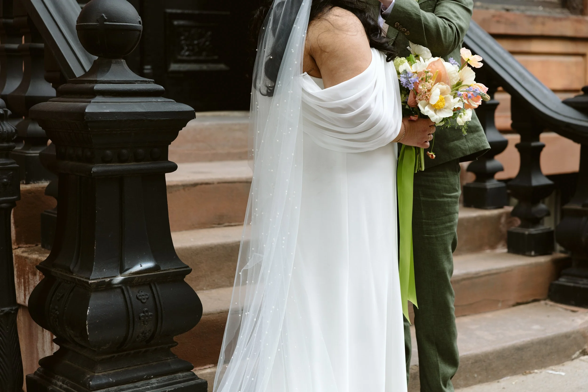 A bride holding a bouquet of flowers, wearing a white wedding dress and veil, next to a person in a green suit, standing on steps outside a building with a black iron fence. marigolds florals nyc florist wedding florals houston hall nyc bar wedding