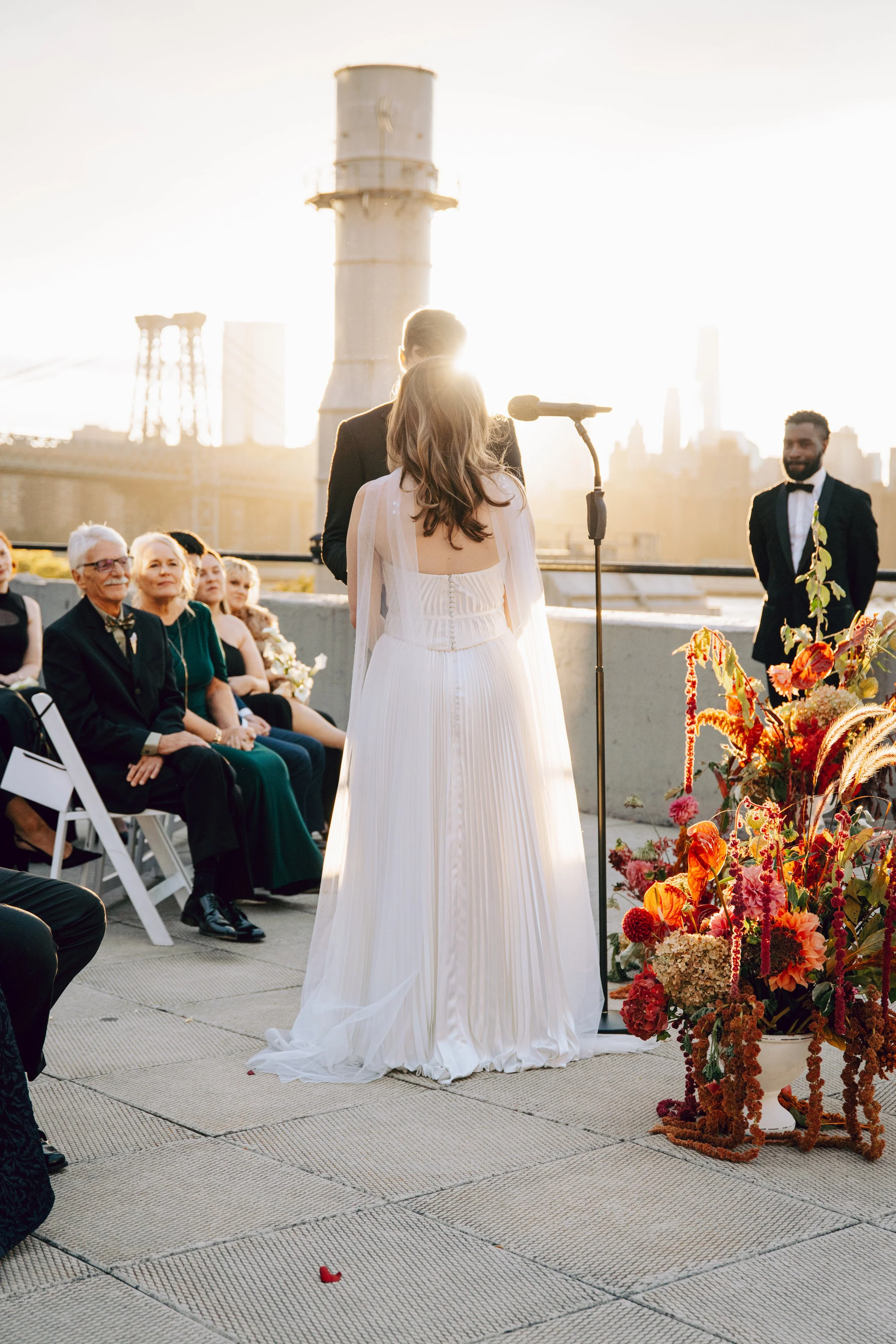 A wedding ceremony on a rooftop with a city skyline, including a large industrial chimney, in the background. The bride and groom's backs are shown, with the bride wearing a white dress and the groom in a dark suit. Guests sit on white chairs, and a 
