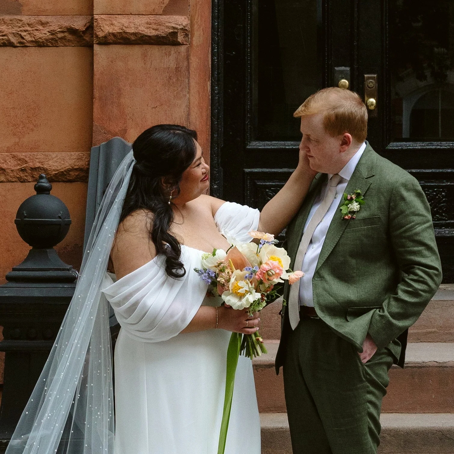 A bride and groom sharing an intimate moment outside a building with a black door. The bride is holding a bouquet of flowers, and they are touching each other's faces tenderly. marigolds florals nyc florist wedding florals houston hall nyc bar weddin