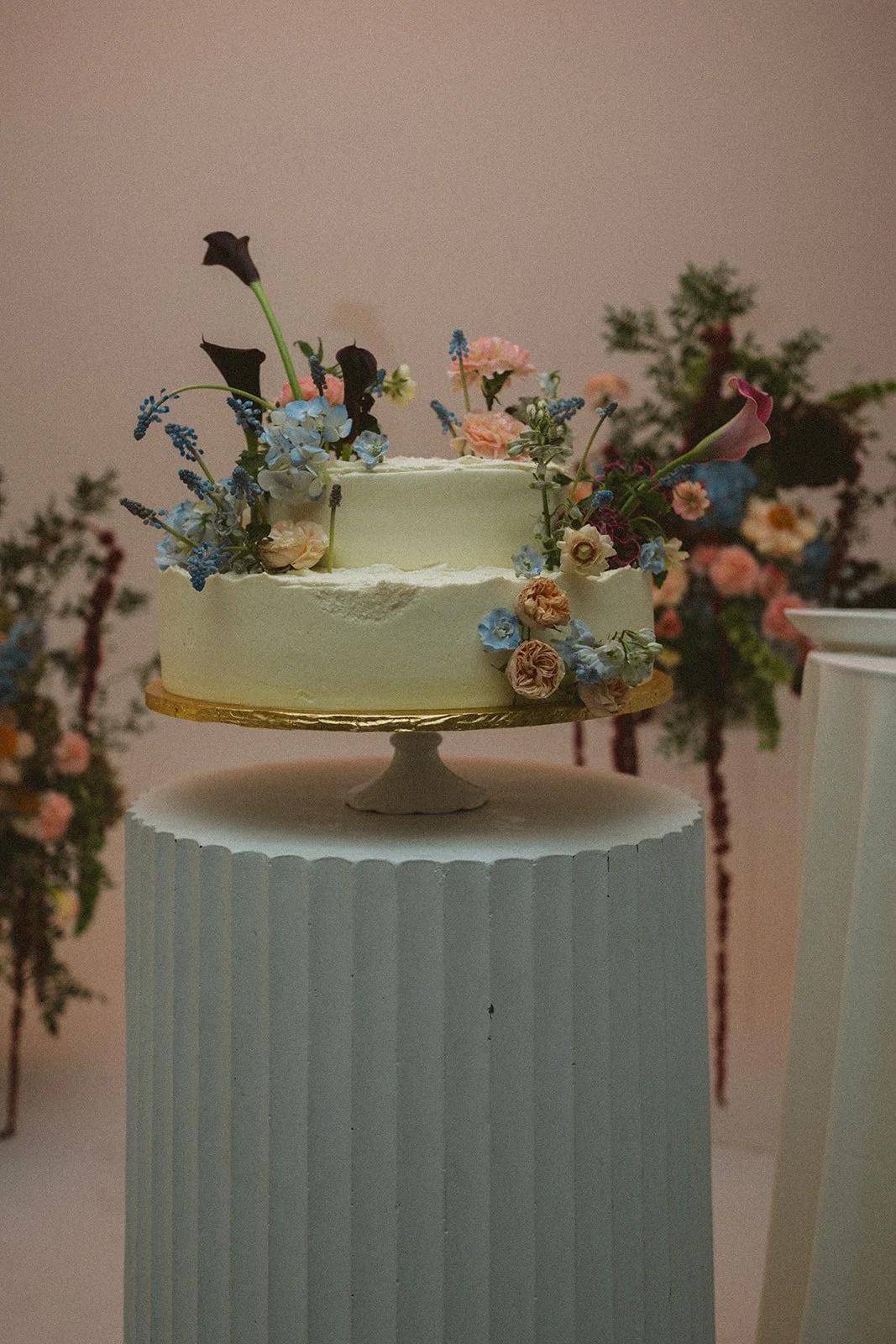 A white wedding cake decorated with pastel-colored flowers and greenery, placed on a gold cake board on a white pedestal, with floral arrangements in the background. marigolds florals nyc florist wedding florals atmosfera wedding