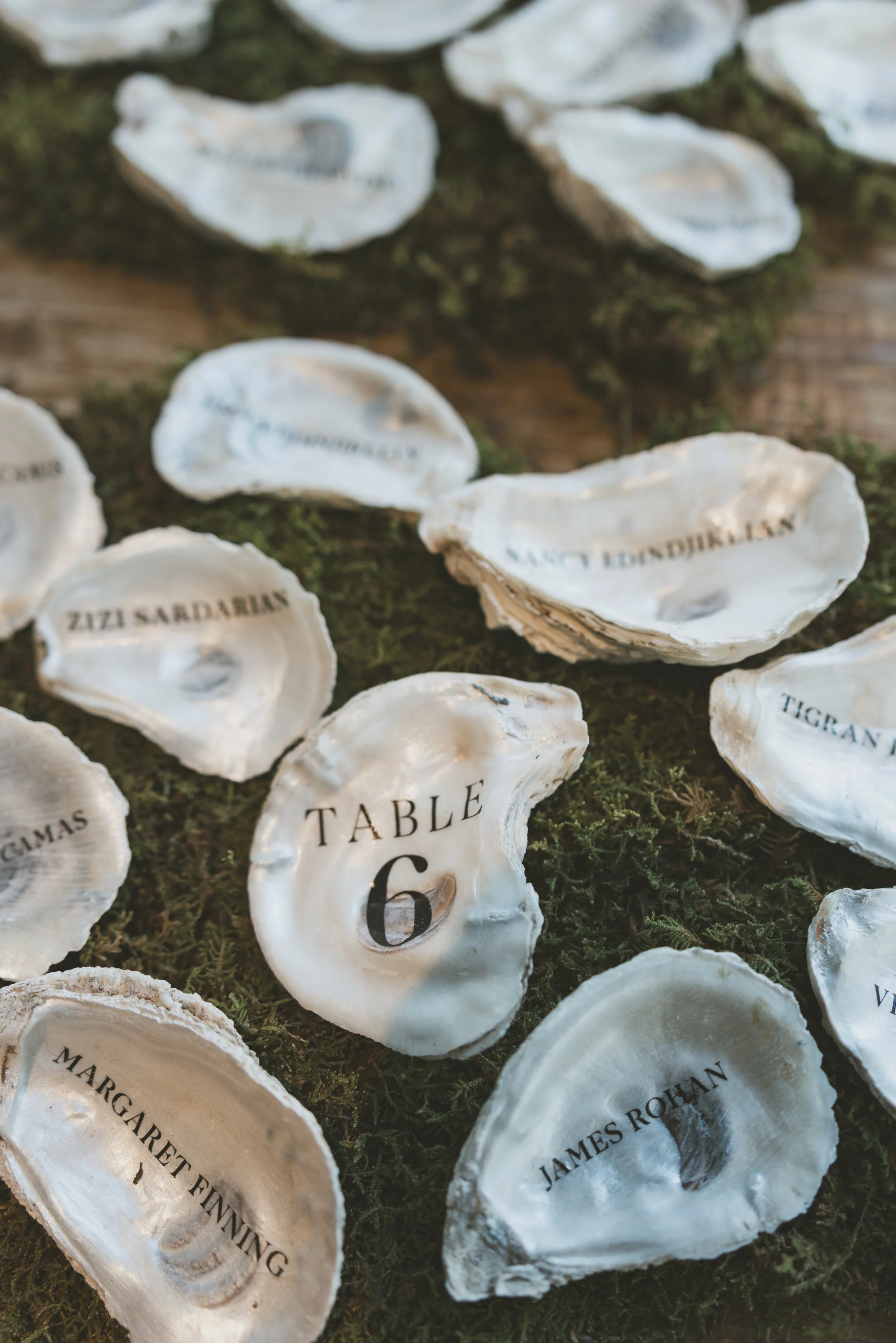 Close-up of oyster shells with names printed on the inside, placed on mossy surface, with some shells labeled 'James Rohan,' 'Margaret Finning,' 'Table 6,' and others.  marigolds florals nyc florist wedding florals the garrison