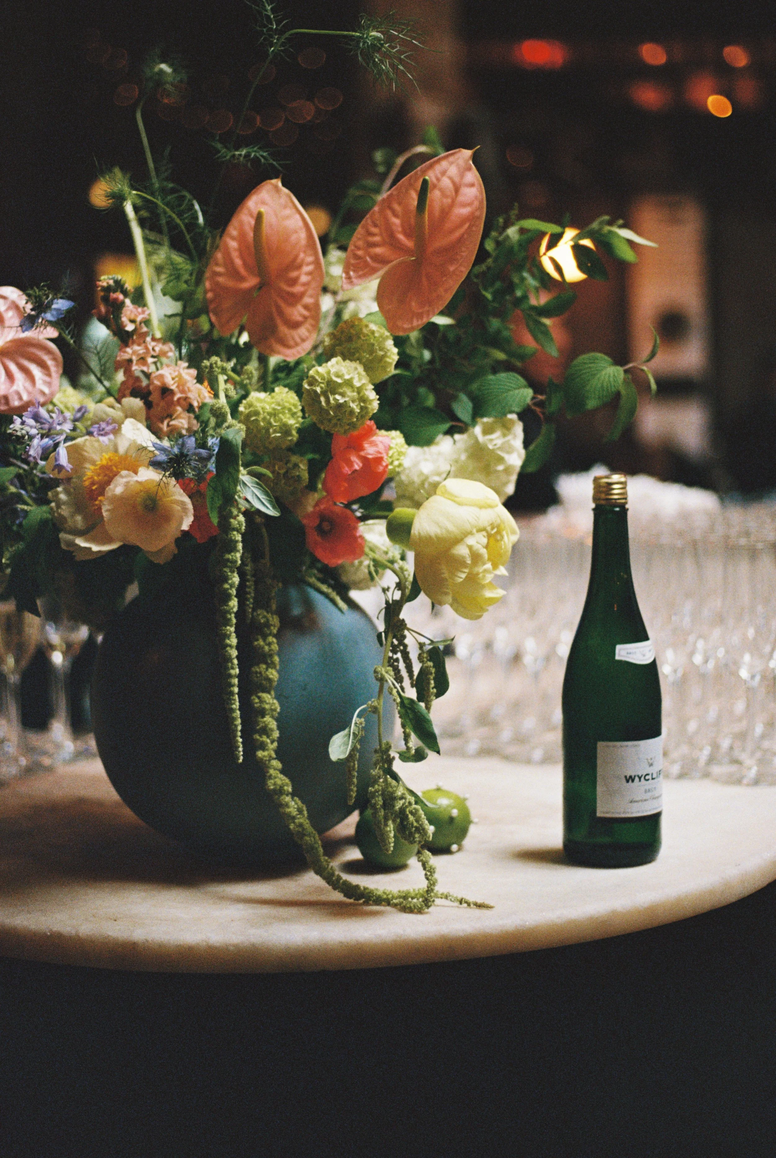 A dark floral arrangement in a black vase with various colorful flowers, green foliage, and hanging green beans, placed on a table. There is a green wine bottle on the table beside the arrangement, with a partially visible white wine label. The backg