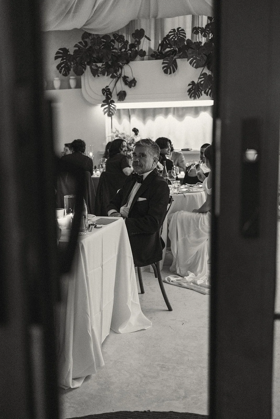 Black and white photo of a formal dinner scene seen through a partially opened door. A man in a tuxedo sits at a table, looking towards the camera. Other guests are visible in the background at similar tables with tableware and glasses, in an elegant