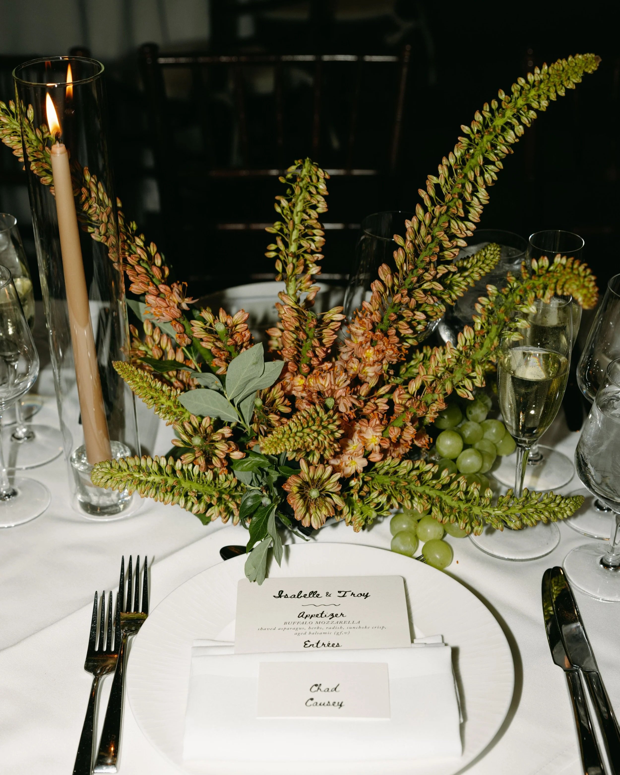A wedding table setting with a floral arrangement of pink and green flowers, several champagne glasses, and place cards for guests, y and a menu describing buffalo mozzarella appetizer and other entrees. marigolds florals