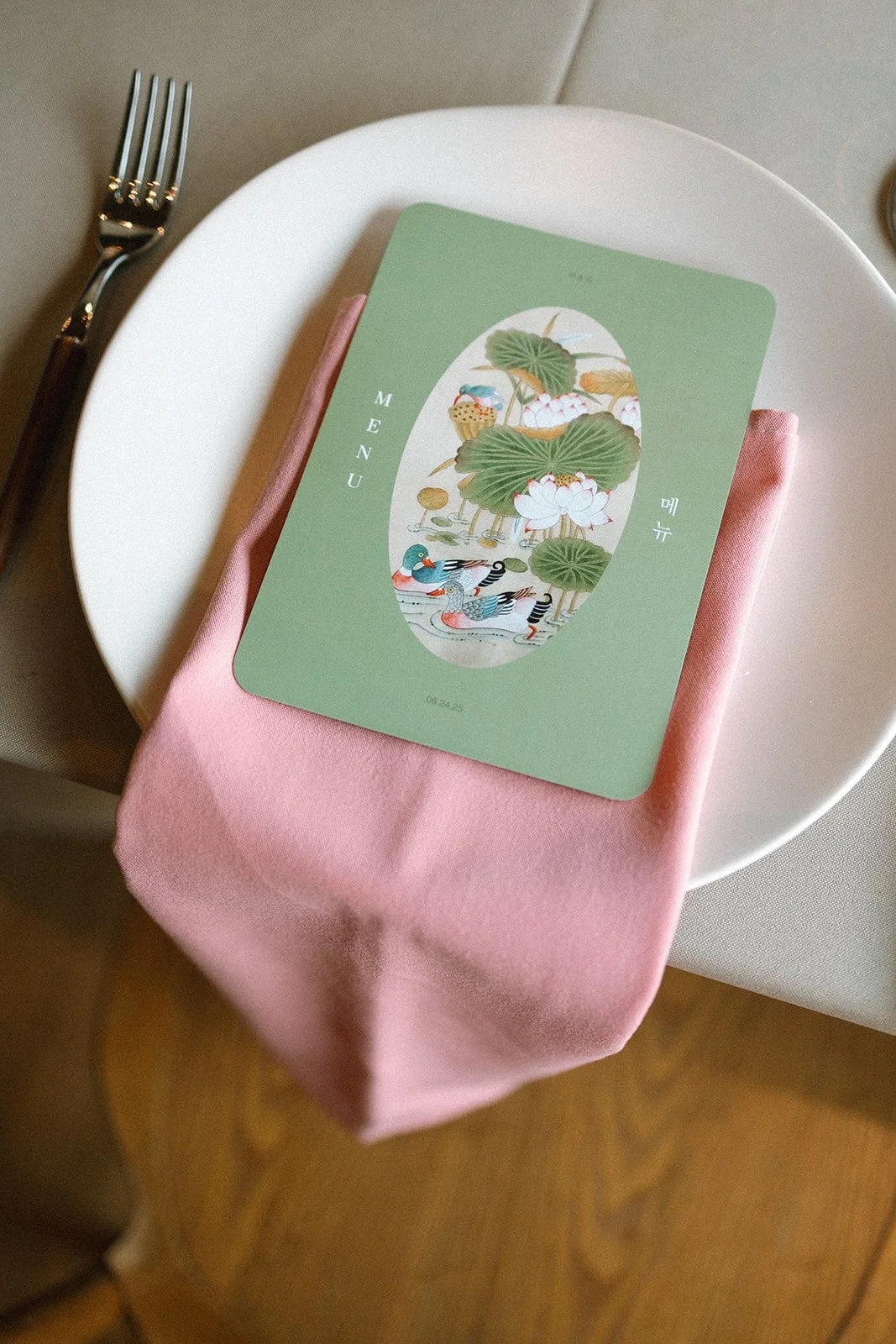 A table setting with a white plate, a pink napkin, and a fork. On the napkin, there is a green menu card with a colorful illustration of ducks, lotus flowers, and lily pads, resembling traditional Asian art. marigolds florals