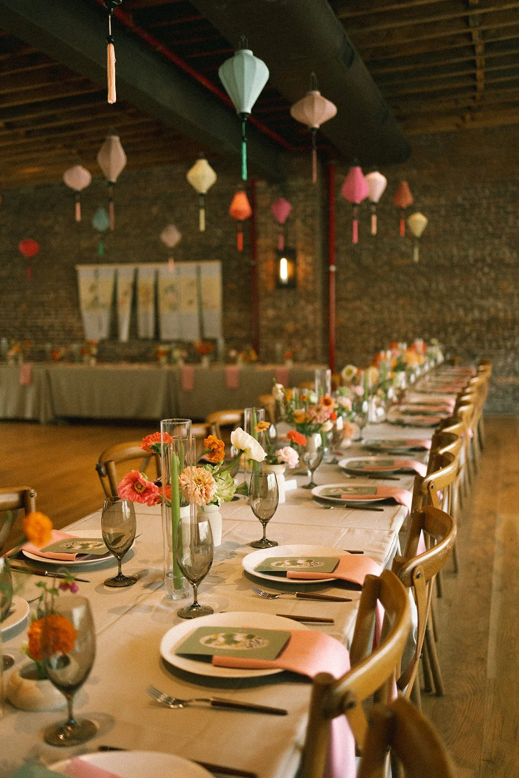 A long dining table decorated with flowers, wine glasses, and plates in a rustic venue with hanging lanterns and a brick wall. marigolds florals