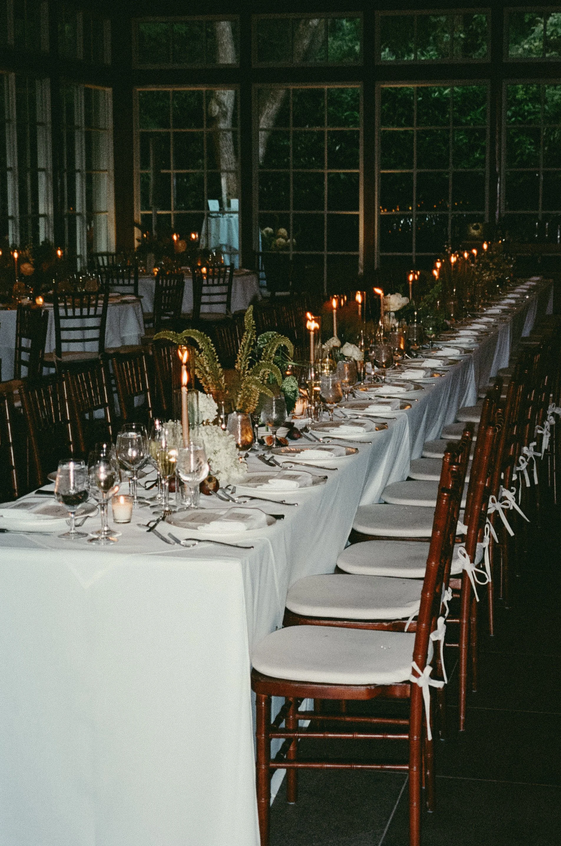 A long dining table with white tablecloths, elegant glassware, plates, silverware, and floral centerpieces, set for a formal event in a dimly lit room with large windows revealing a wooded outdoor scene. marigolds florals nyc florist wedding florals 