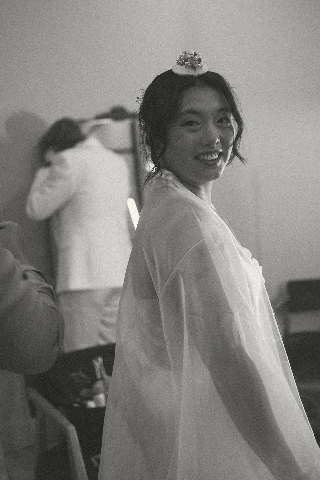 A smiling woman with dark hair styled in loose waves, wearing a decorative headpiece, turns her head to smile at the camera. In the background, another woman is seen adjusting her hair, with people and objects faintly visible in a behind-the-scenes s